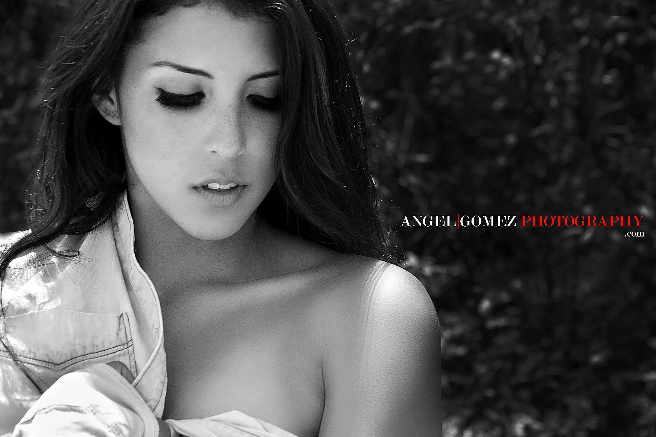 Black and white portrait of a young woman with long dark hair, closed eyes, and freckles, looking down with a slight smile. She is wearing an open-collared shirt, and a strap from her top is visible on her shoulder. The background is blurred foliage.