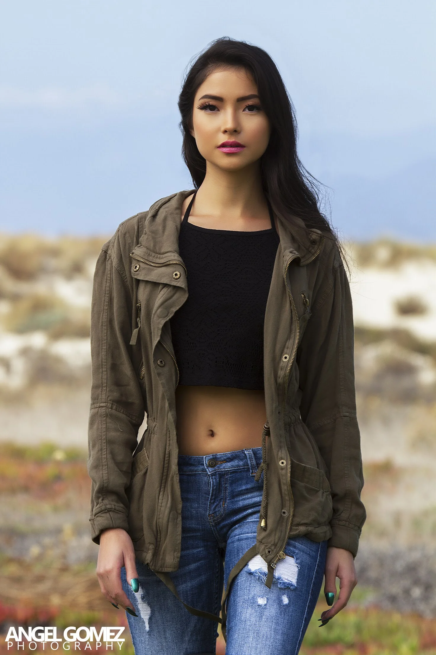 Young woman with dark hair wearing a black crop top, olive green jacket, and ripped blue jeans standing outdoors with a blurred natural background.