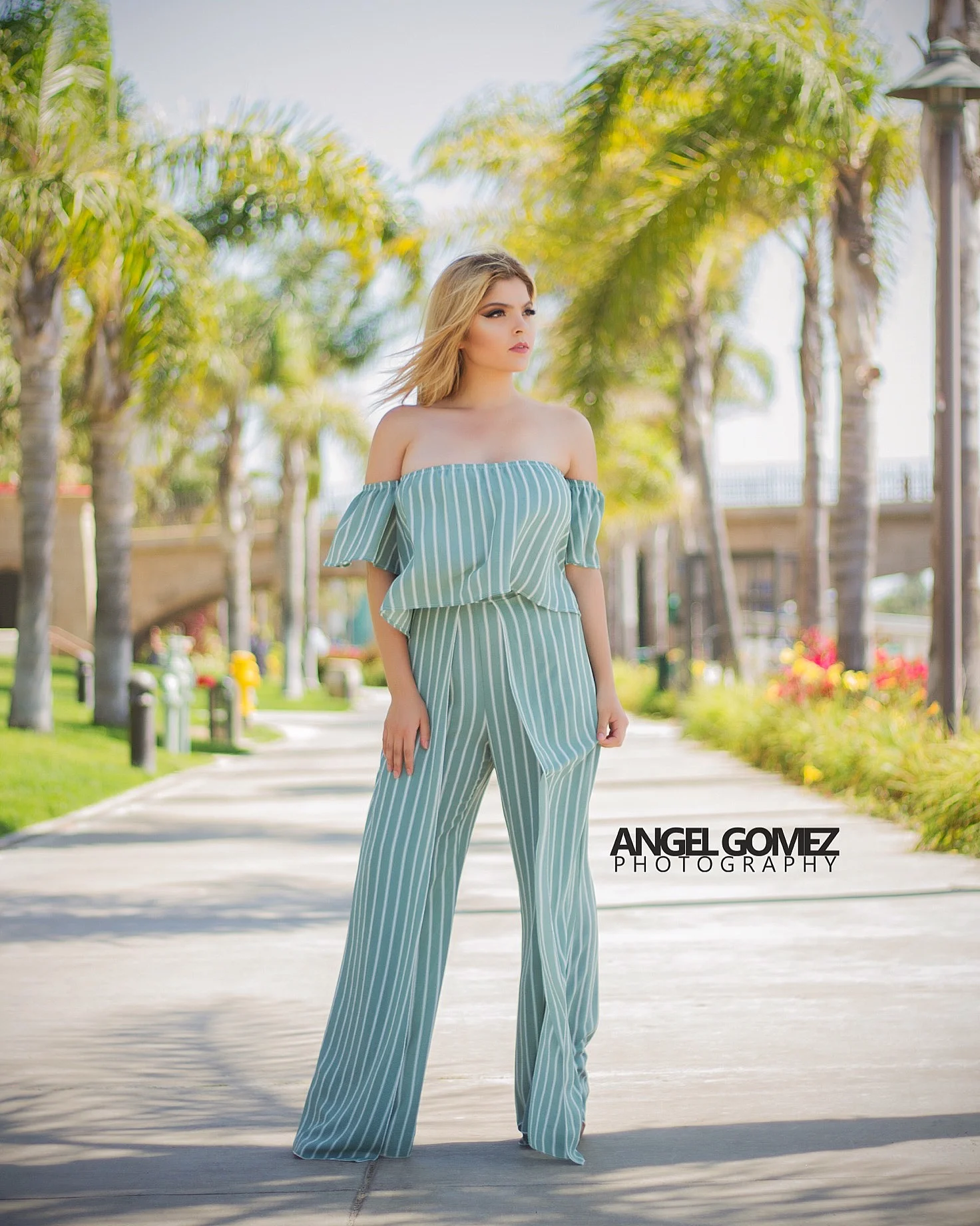 Young woman in a green striped off-shoulder jumpsuit standing on a sunny sidewalk lined with palm trees.