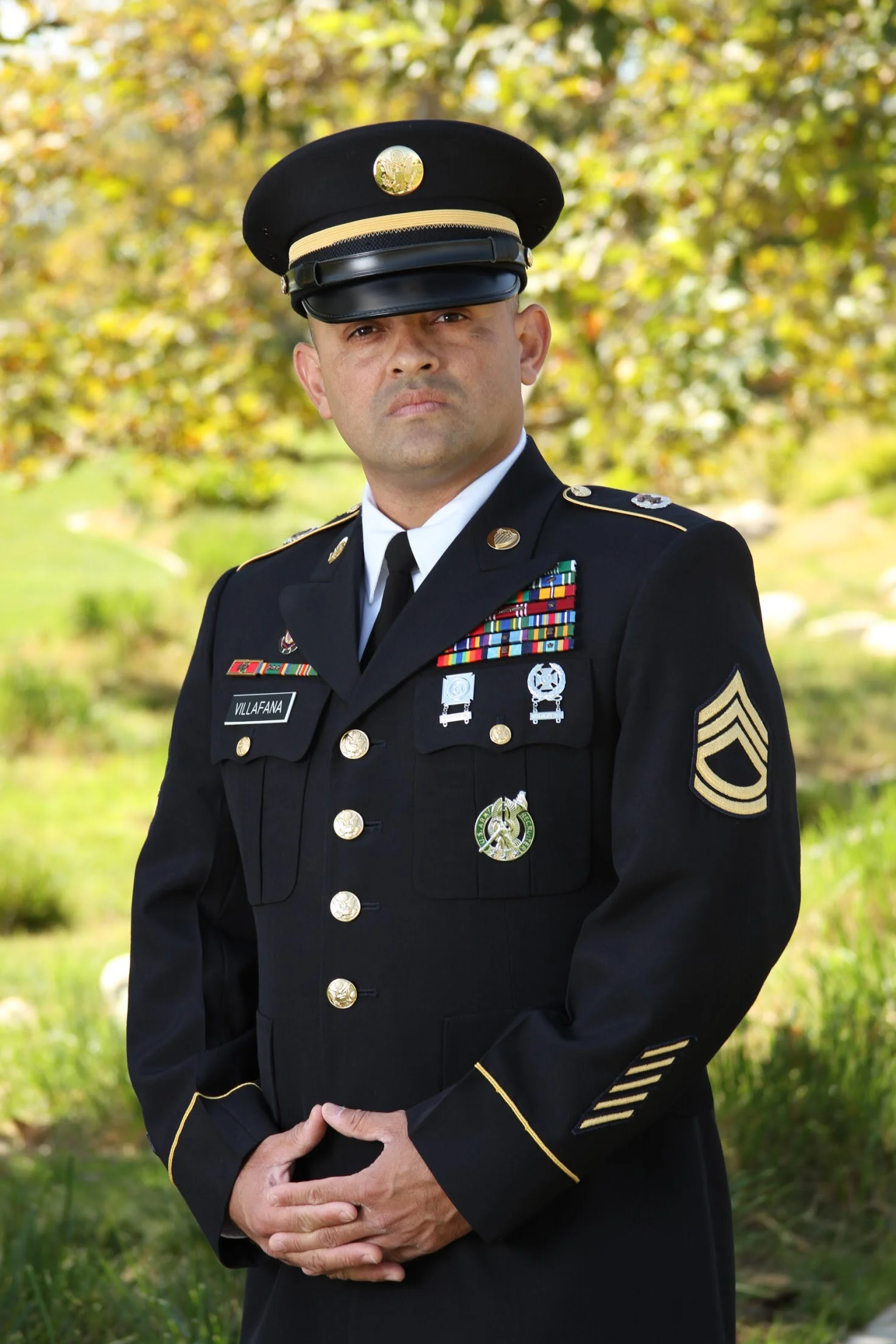 A man in a formal military uniform standing outdoors with trees and greenery in the background.