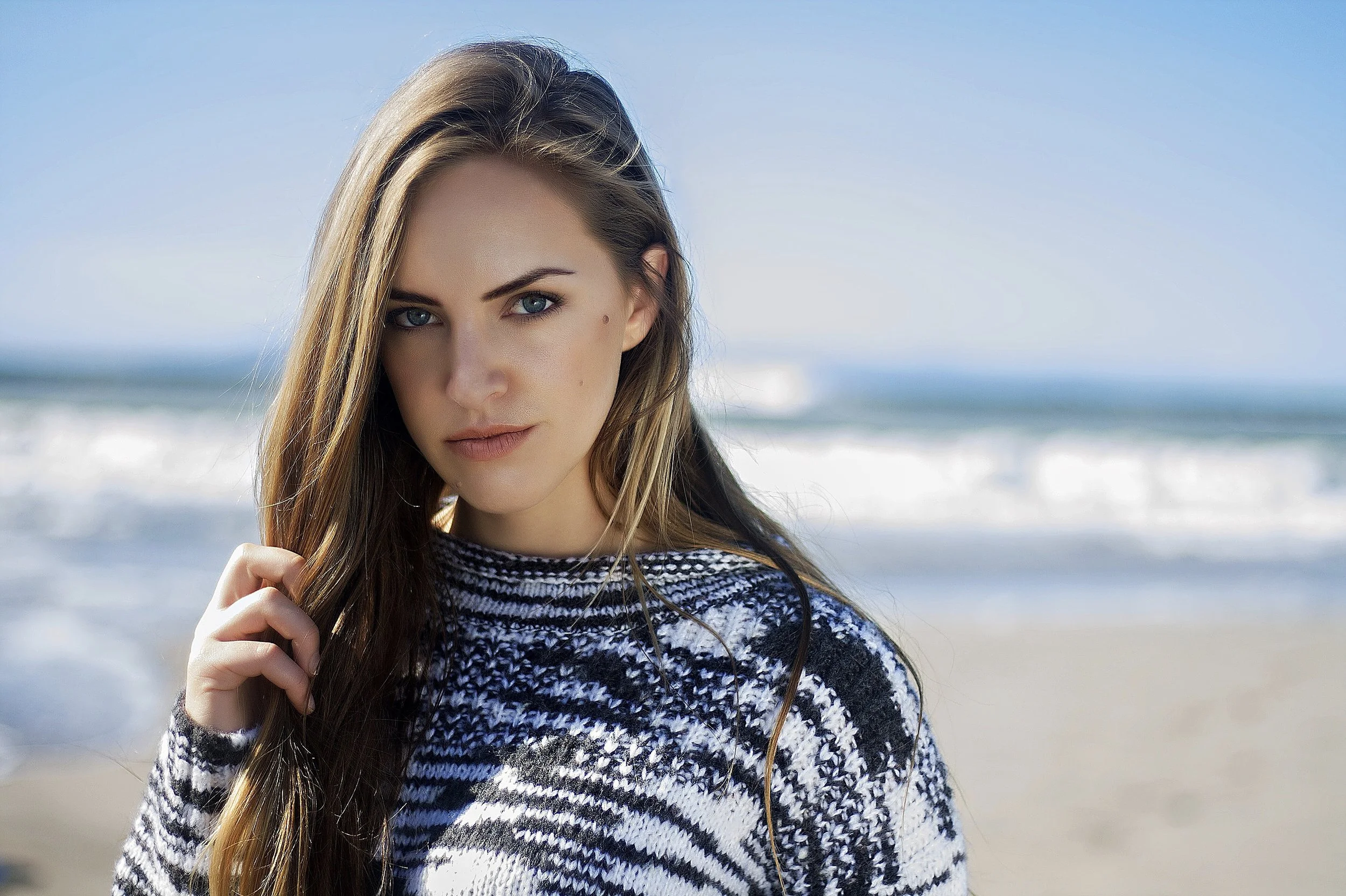 A woman with long brown hair and blue eyes standing on a beach with waves in the background, wearing a black and white patterned sweater.