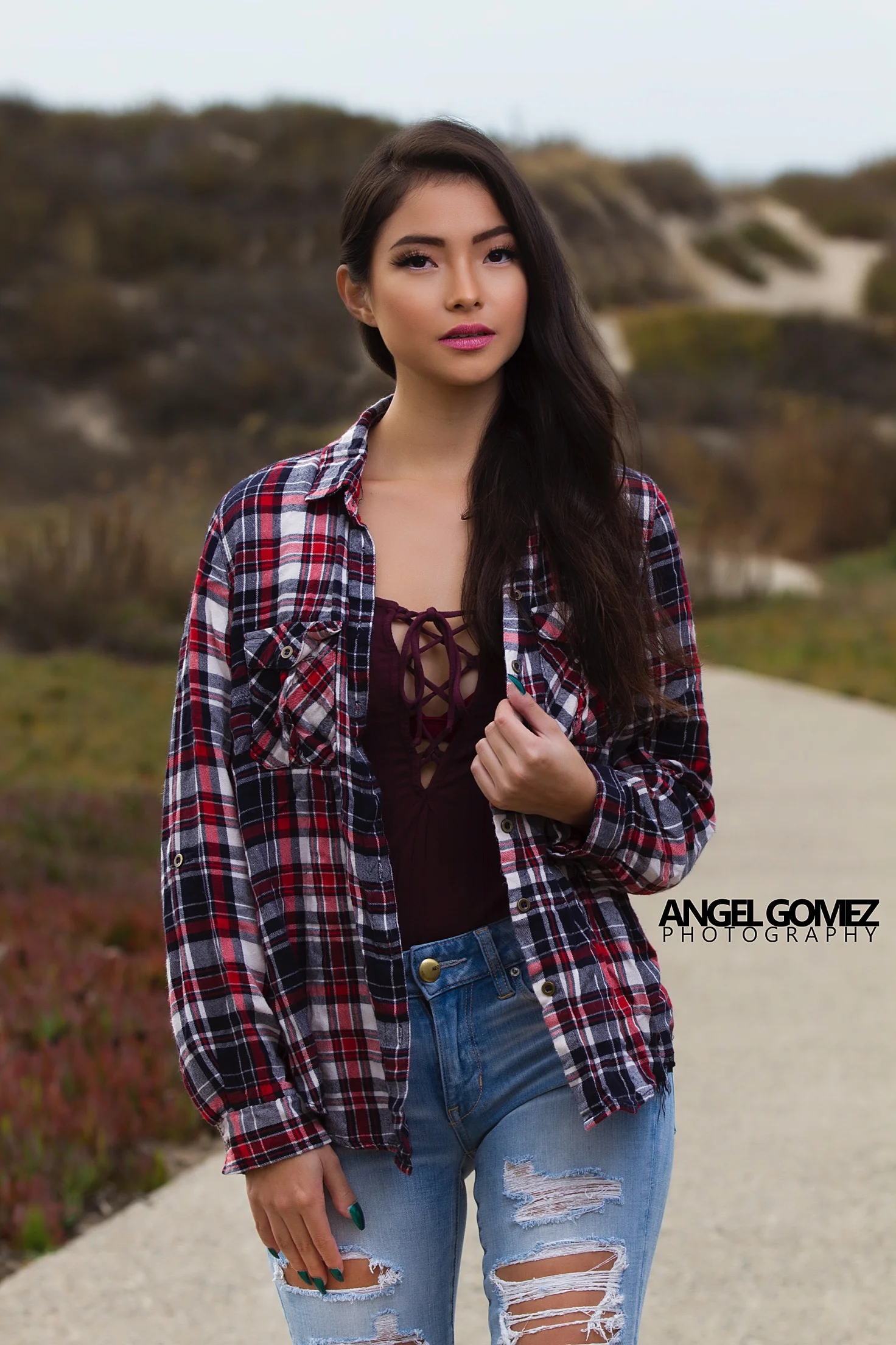 A young woman with long dark hair standing outdoors on a pathway, wearing a plaid shirt over a lace-up top and distressed jeans, with a blurred natural landscape in the background.