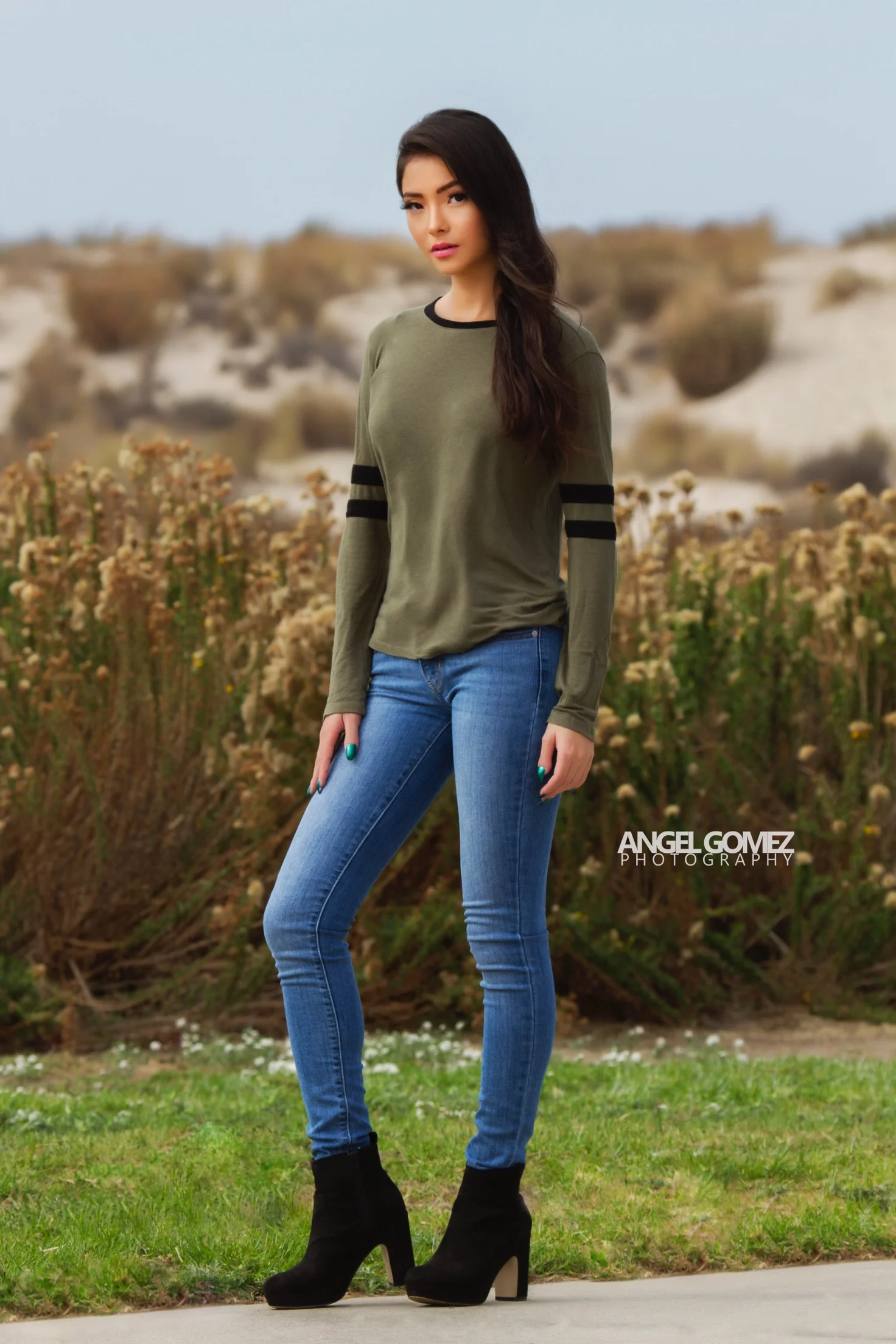 A young woman with long dark hair, wearing an olive green long-sleeve shirt with black stripes on the sleeves, blue jeans, and black high-heeled boots, standing outdoors in a natural setting with grass and shrubbery.