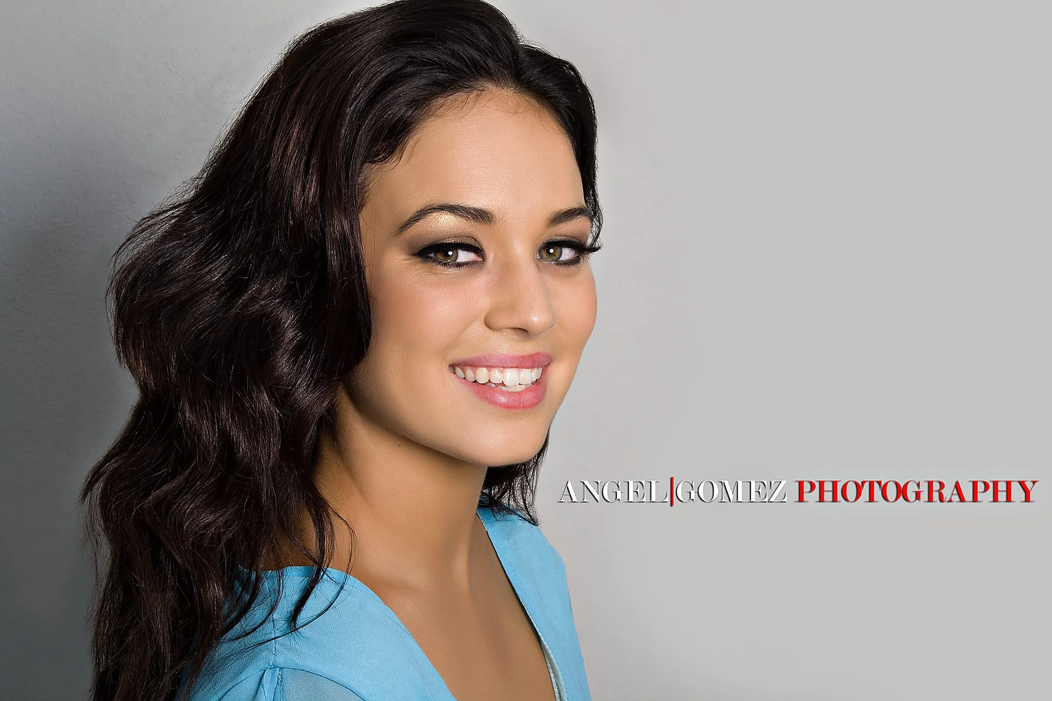 Portrait of a young woman with dark wavy hair, light makeup, and a blue top smiling against a gray background with "Angel Gomez Photography" logo.