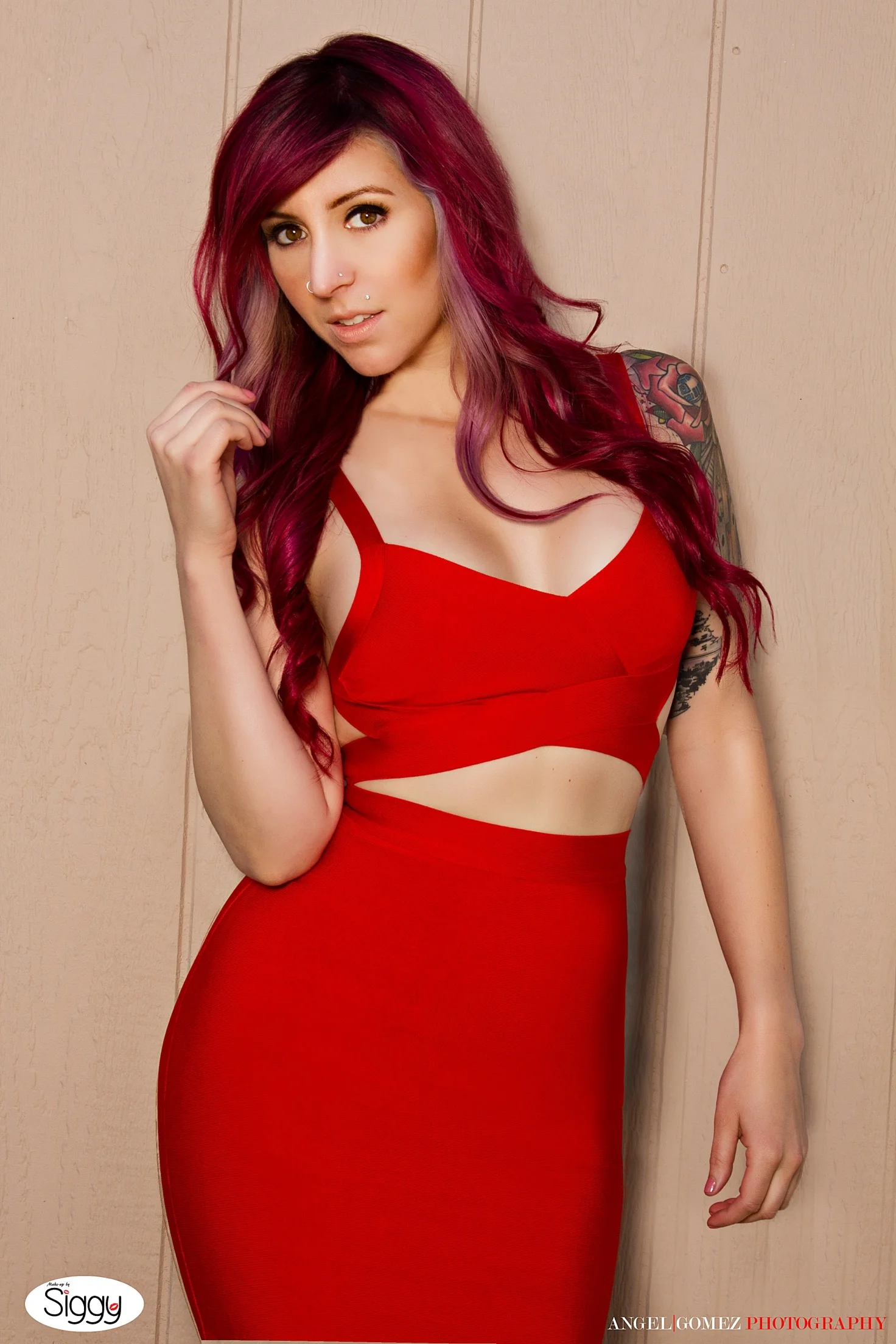 A woman with long, wavy, red hair and tattoos on her right arm, wearing a red cut-out dress, standing against a beige wall.
