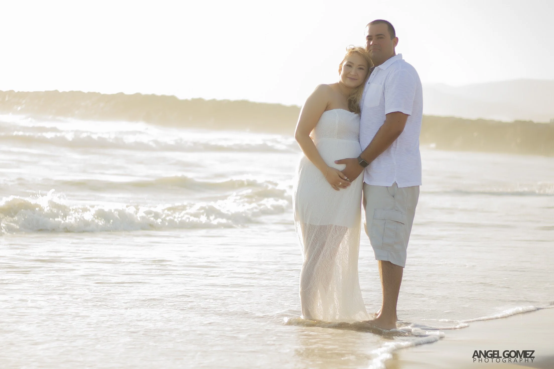 A pregnant woman and a man standing together on the beach during sunset, both smiling, with waves in the background.