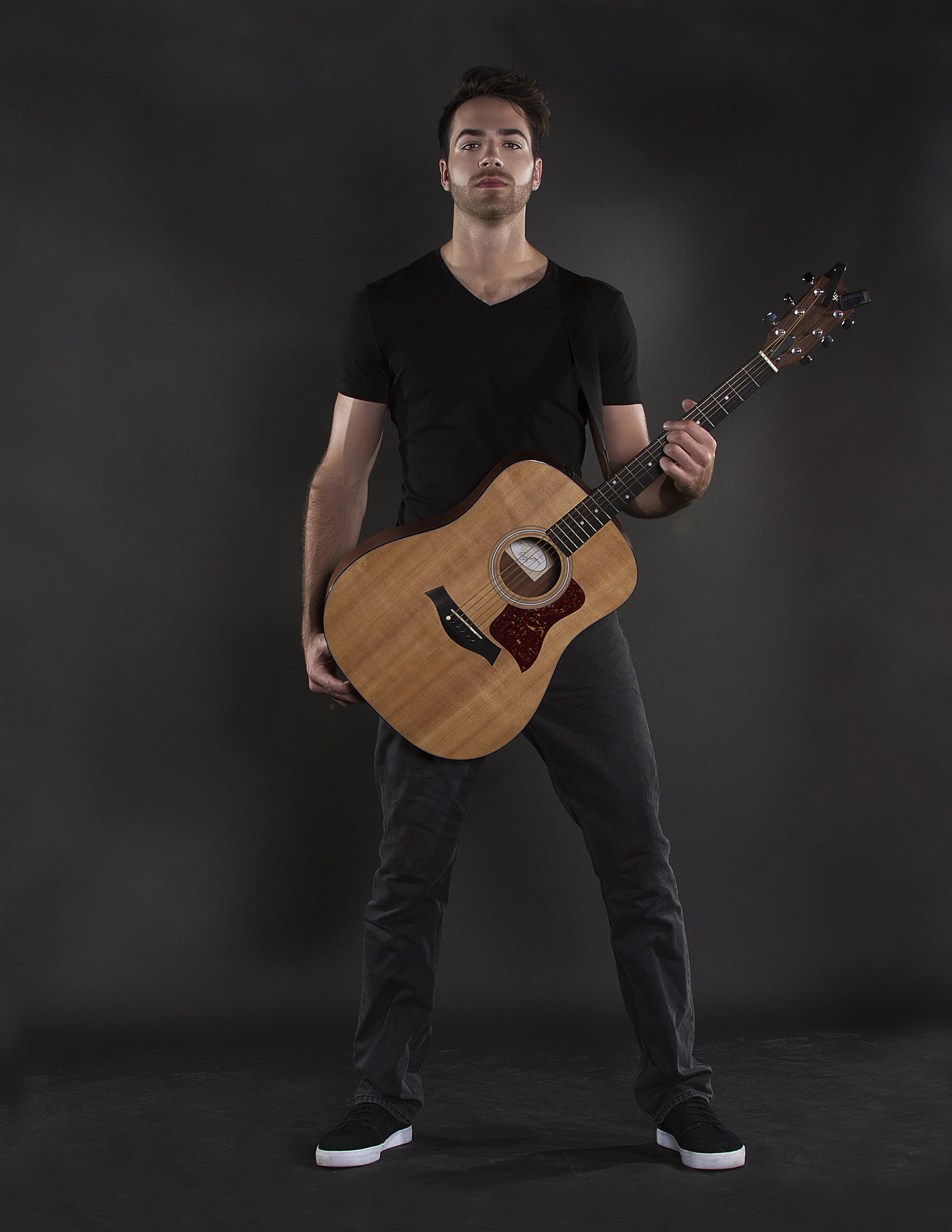 A young man in black t-shirt and dark pants holding an acoustic guitar against a dark background.