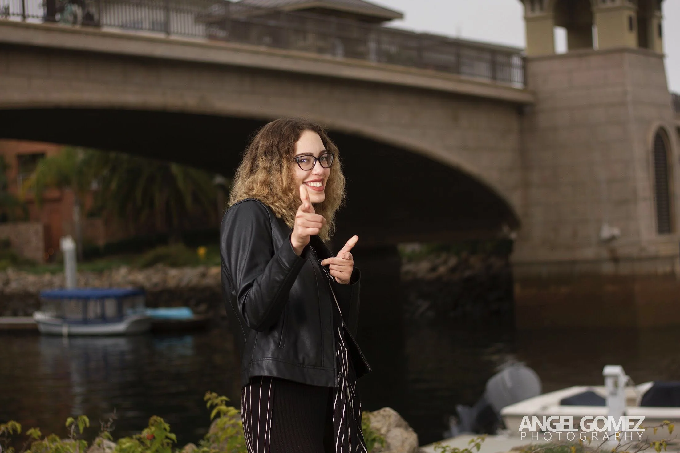 A woman with curly hair and glasses is smiling and pointing at the camera, standing near a body of water with boats and a large bridge in the background.