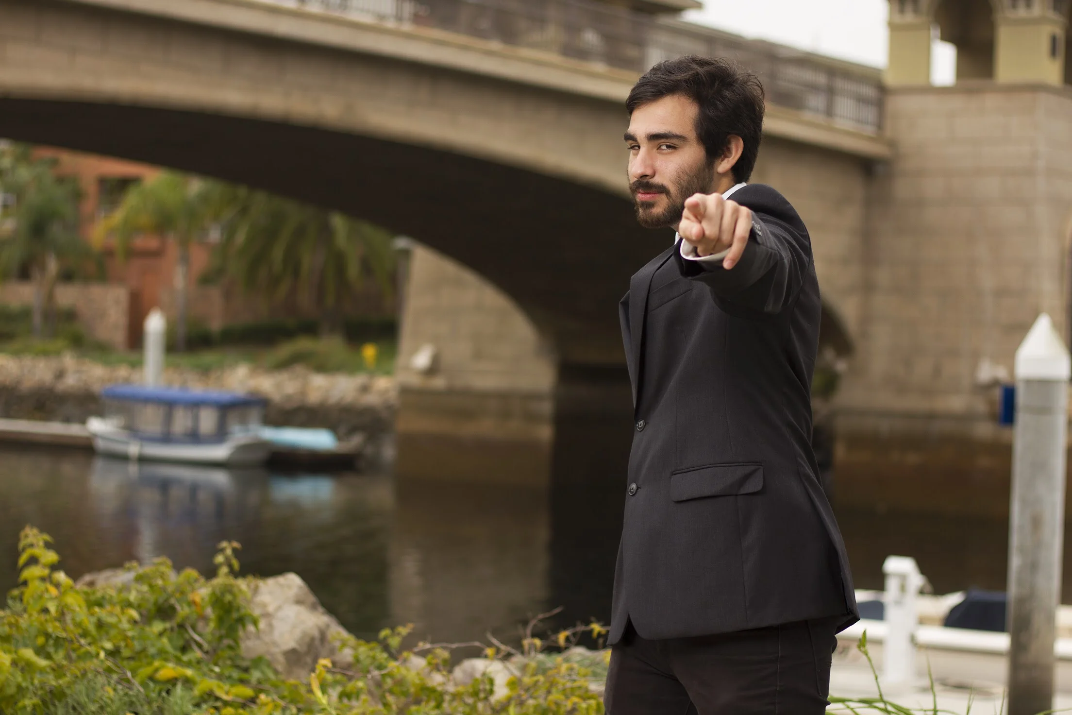 A man in a black suit pointing towards the camera by a waterway with a bridge and boats in the background.