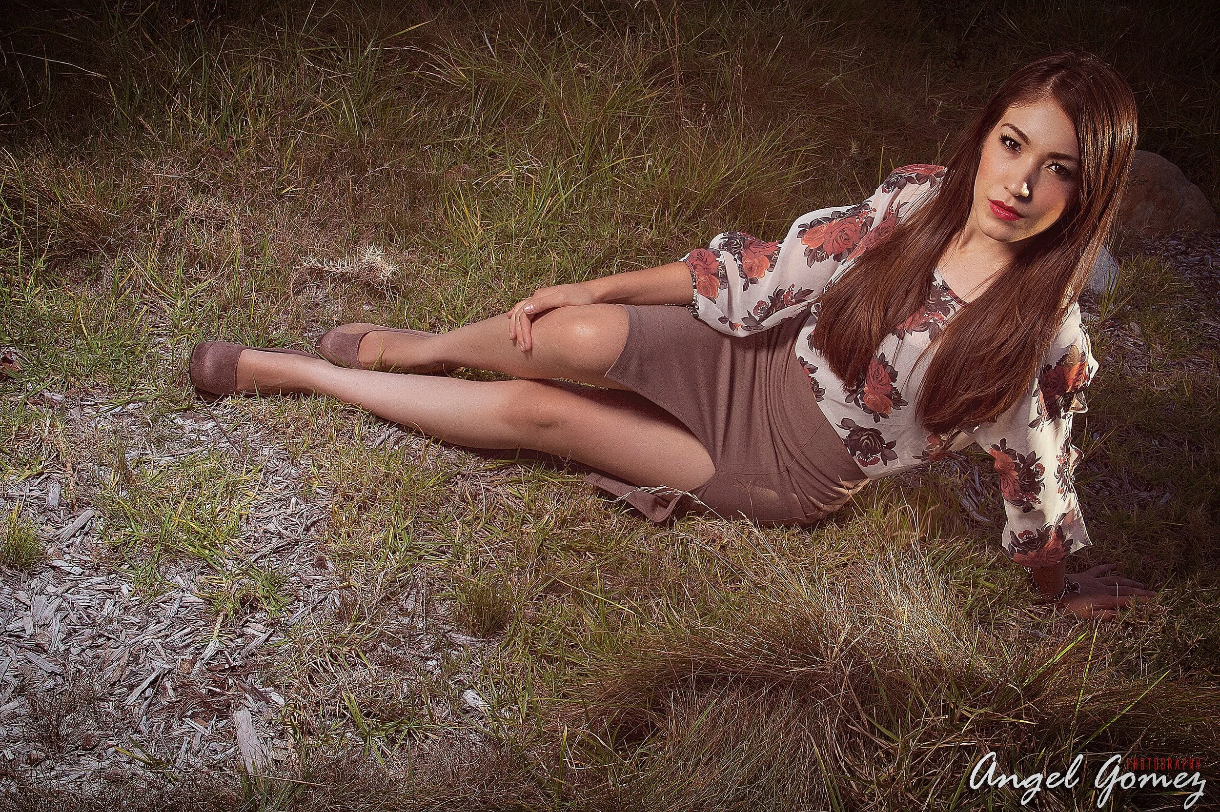 A woman with long brown hair, wearing a floral blouse, beige skirt, and beige shoes, lying on her side on a grassy area at dusk, posing for a photo.