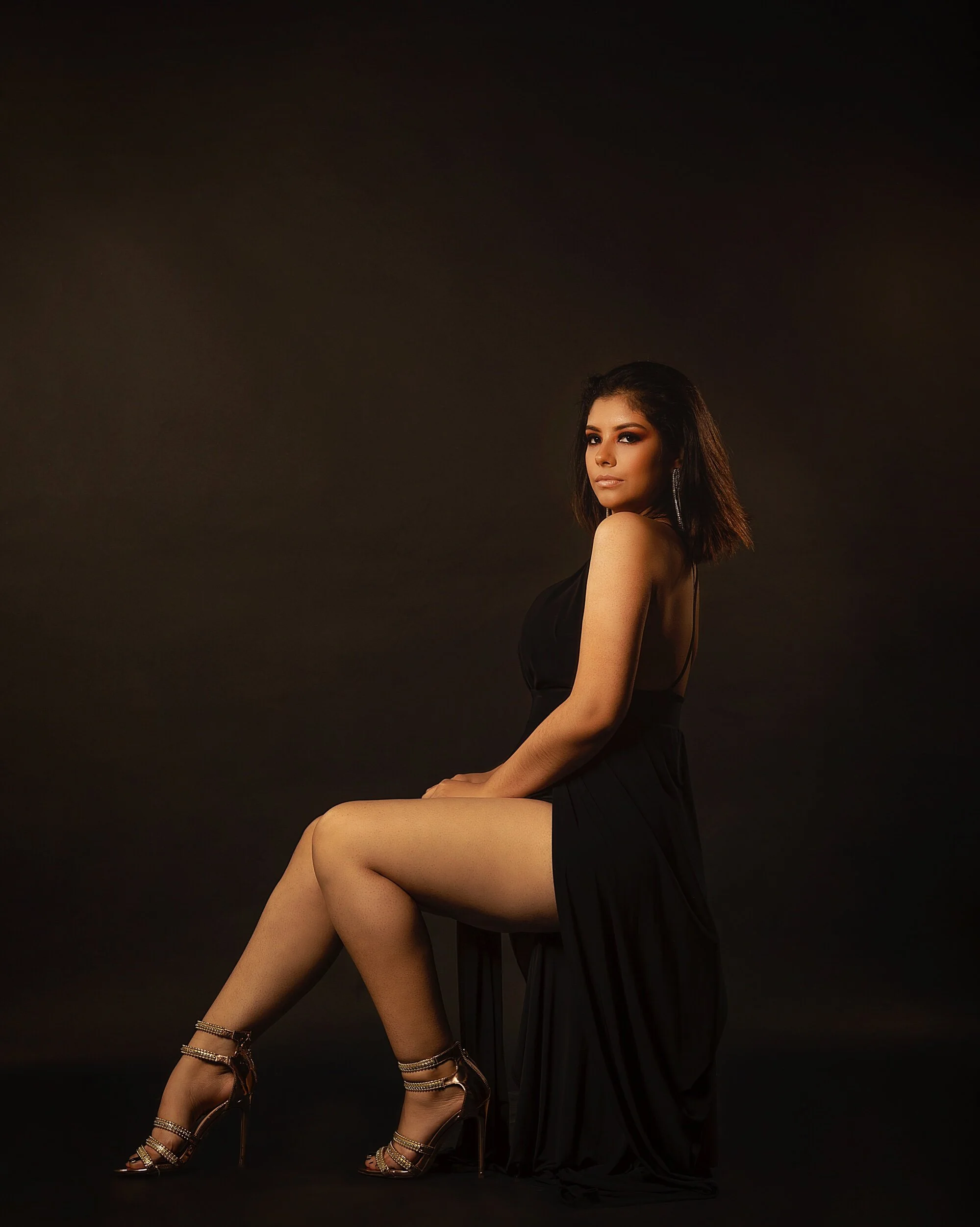 Woman wearing black dress and high heels sitting against a dark background.