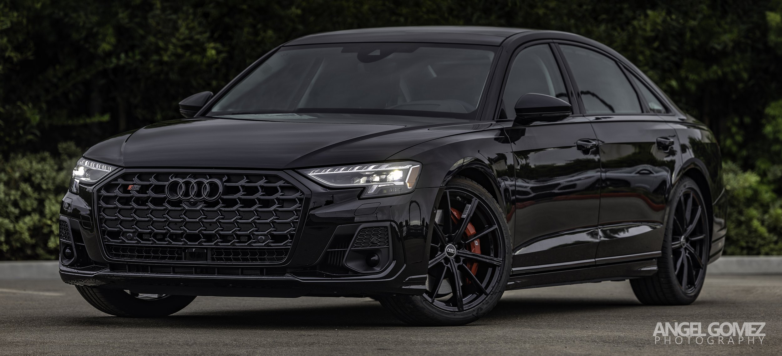 Black Audi sedan with red brake calipers parked on pavement with green bushes in background.