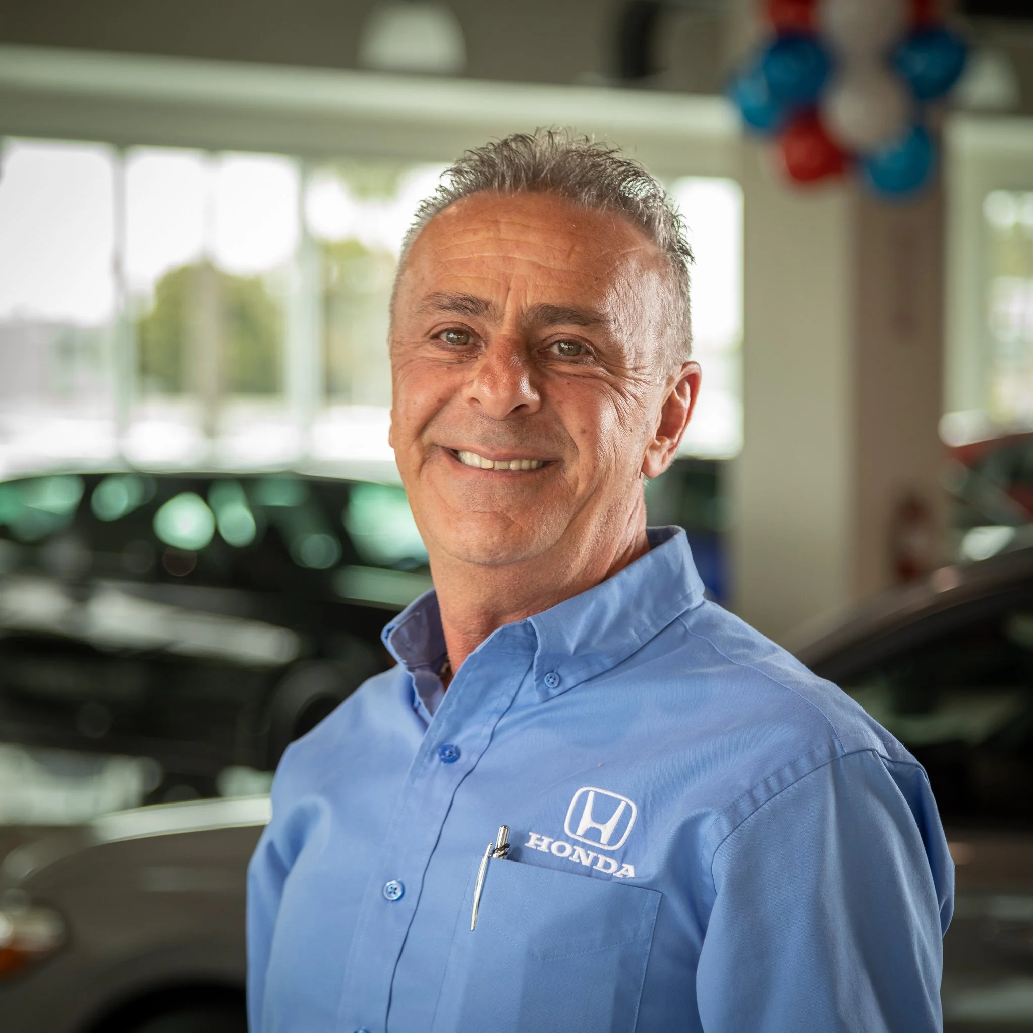 A smiling man with gray hair in a blue hear dealership shirt with Honda logo, standing indoors with cars and balloons in the background.