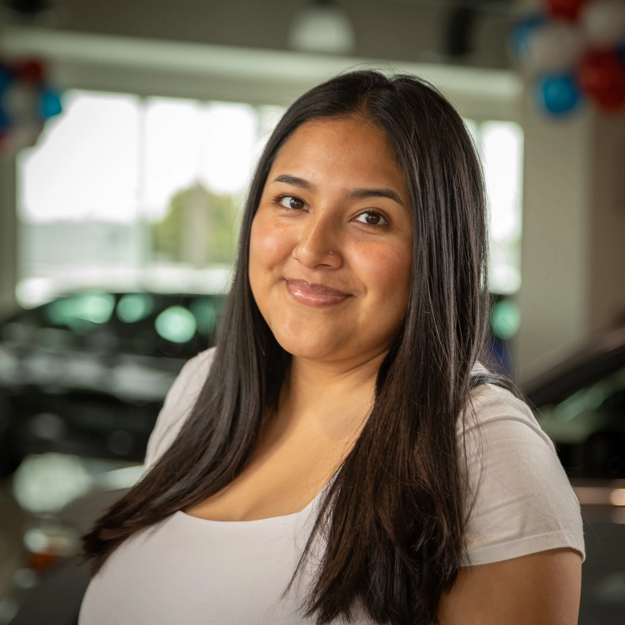 A smiling woman with long dark hair, wearing a white top, standing inside a car dealership.