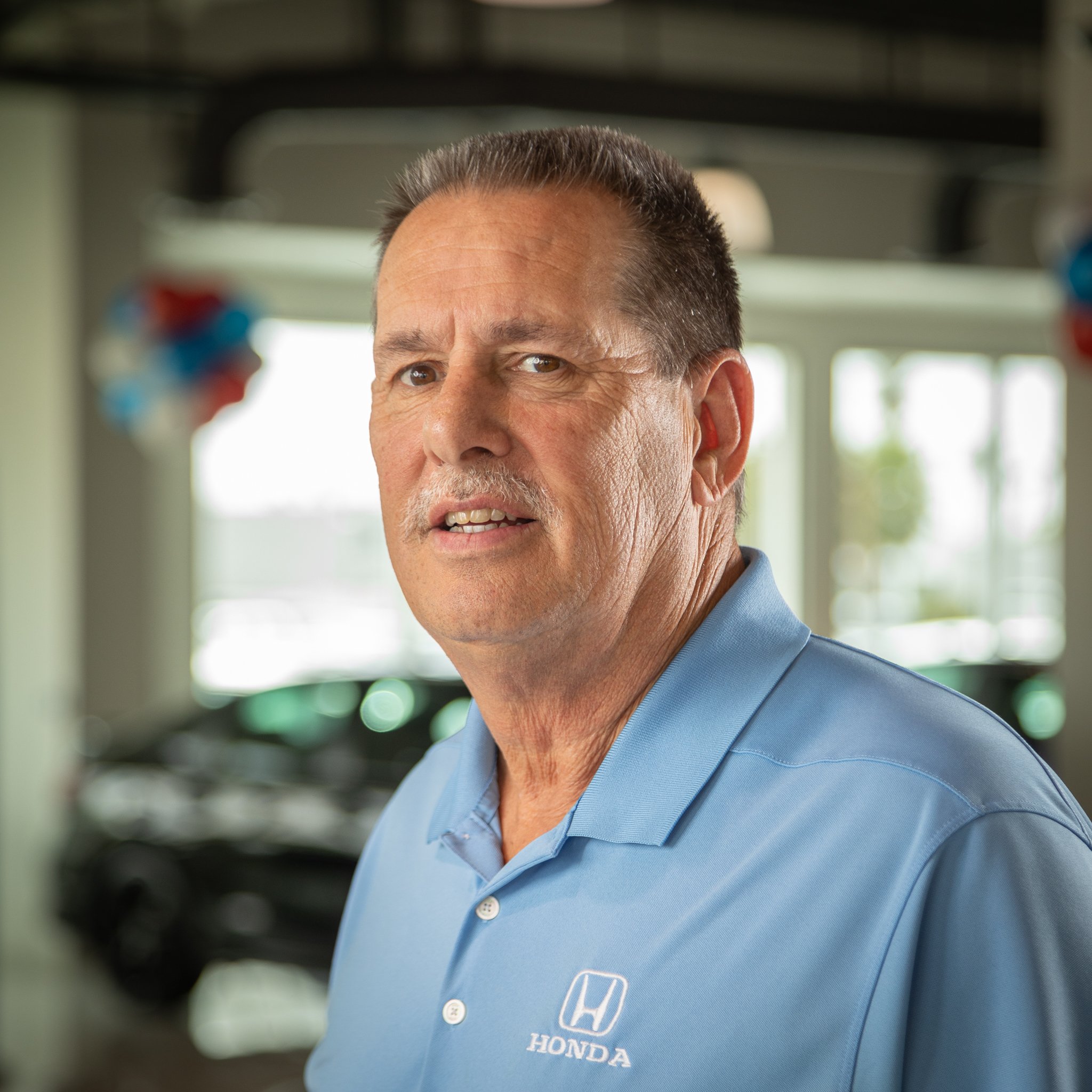 A middle-aged man with short, dark hair and a mustache wearing a light blue collared shirt with a Honda logo, standing inside a Honda dealership.