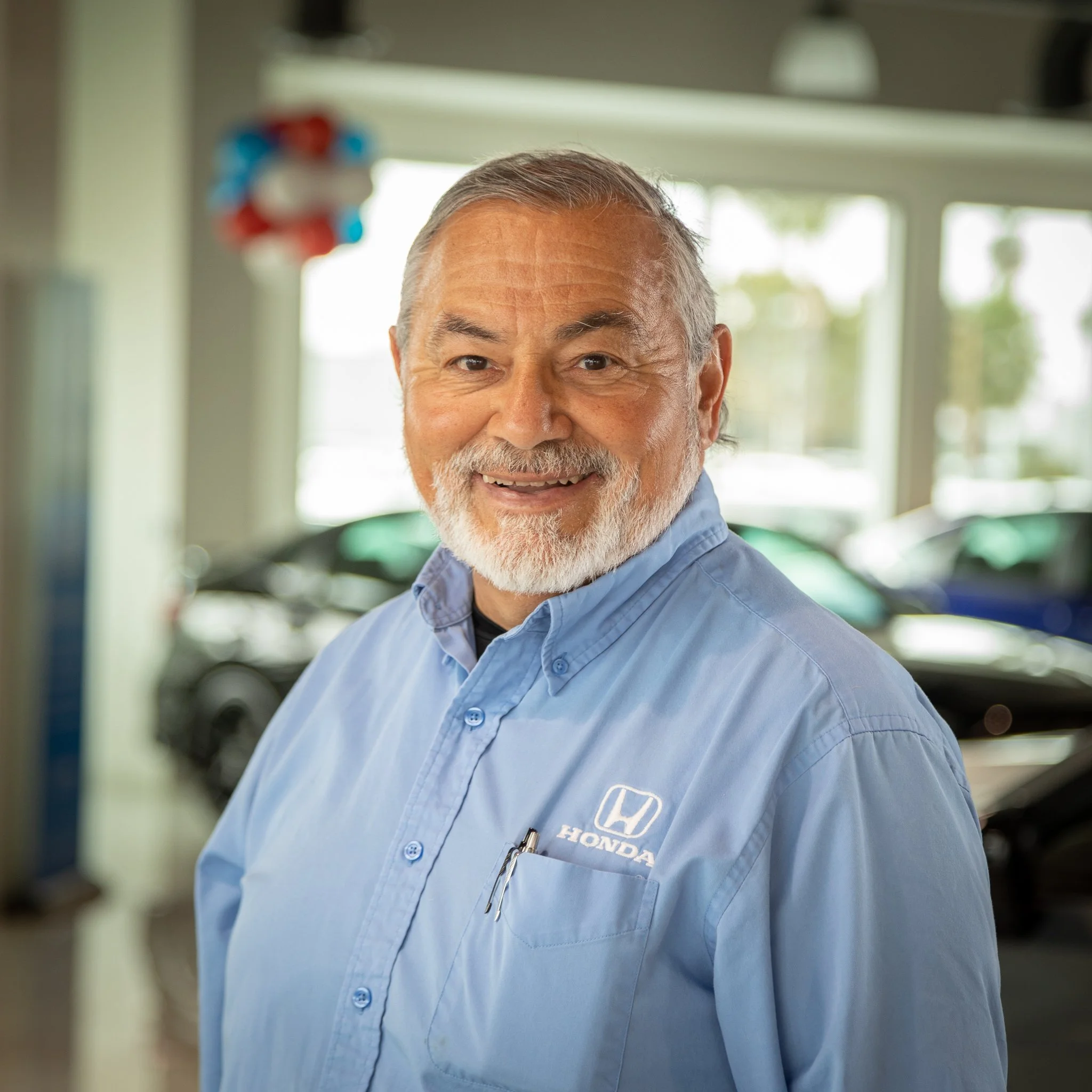 A smiling older man with a beard and gray hair, wearing a blue Honda uniform shirt with a pen in the pocket, standing inside a Honda dealership with cars and windows in the background.