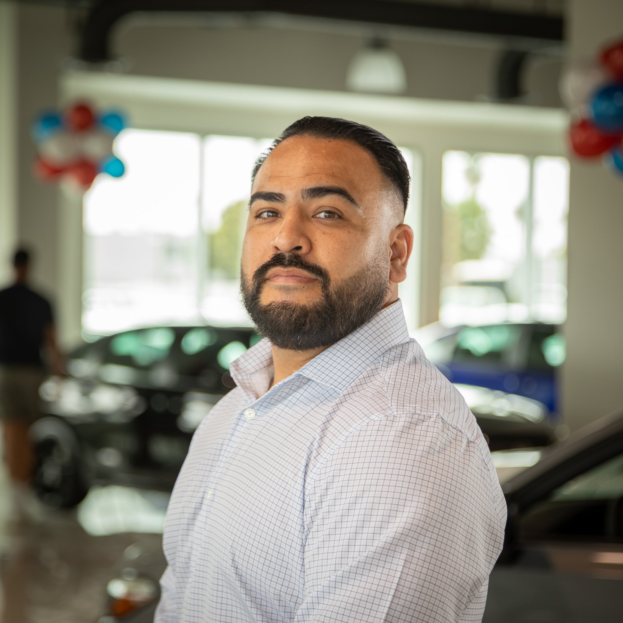 A man with a beard and short dark hair posing inside a car dealership, wearing a light-colored, checkered shirt, with cars and dealership balloons in the background.