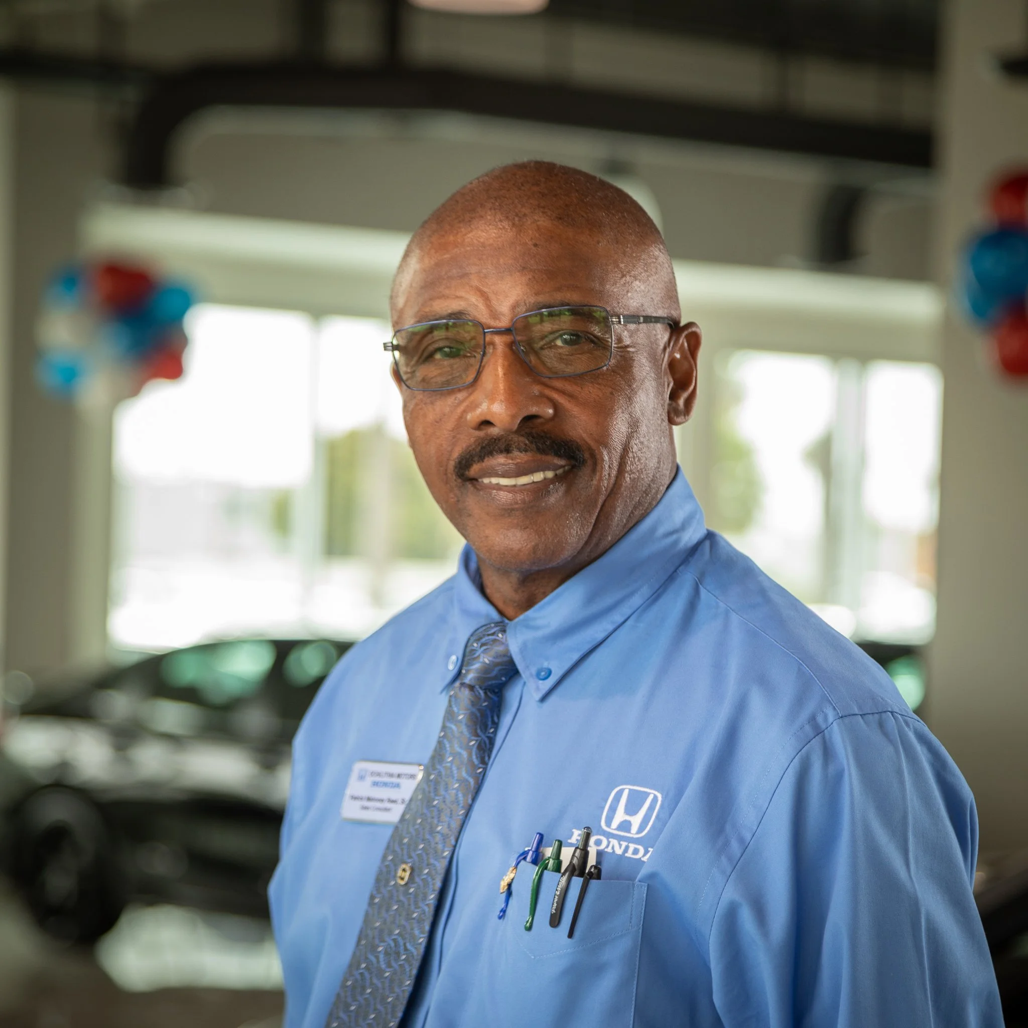 A middle-aged man with glasses wearing a light blue Honda shirt and a patterned tie, standing in a car dealership or service center. He has a friendly expression and appears professional.