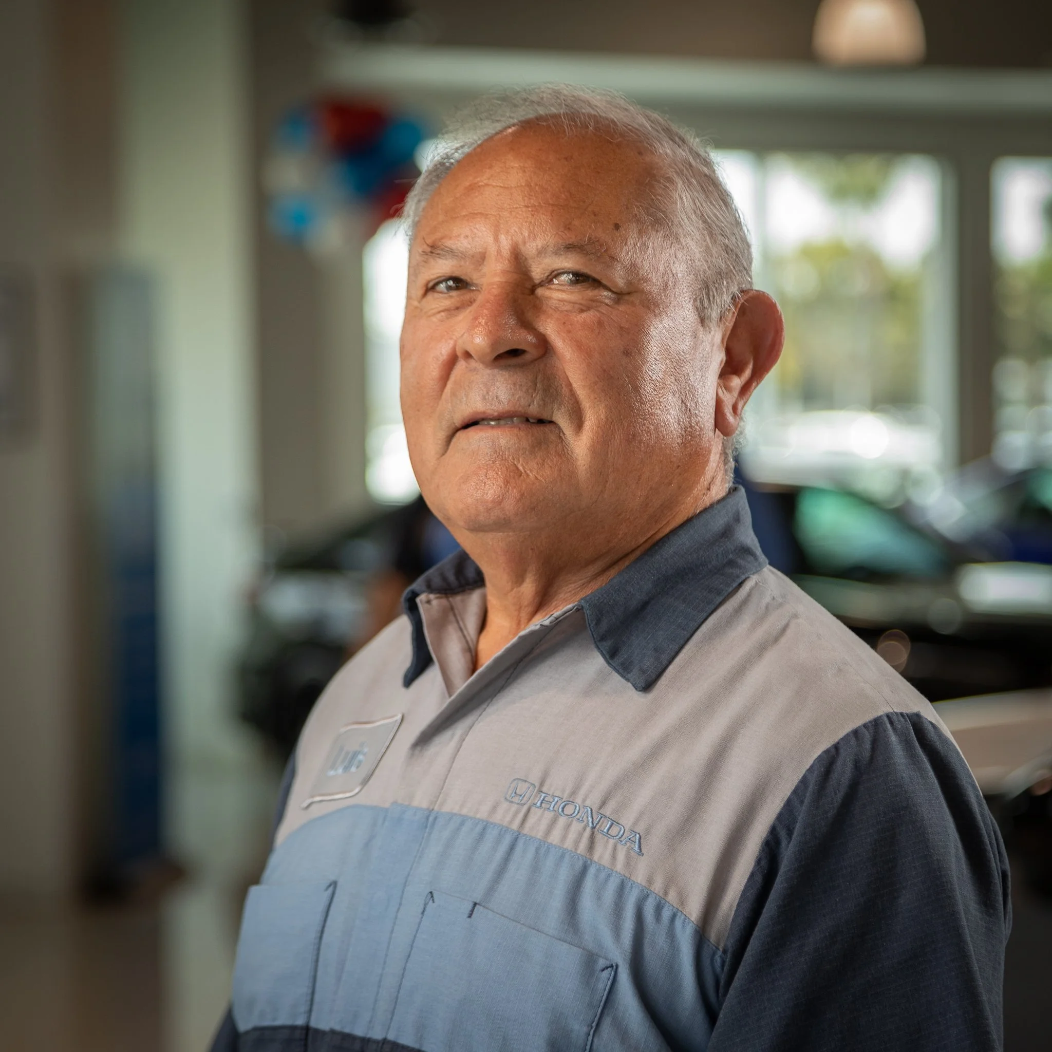 Close-up of an older male car dealership employee in uniform standing inside a dealership, with cars and window view in the background.