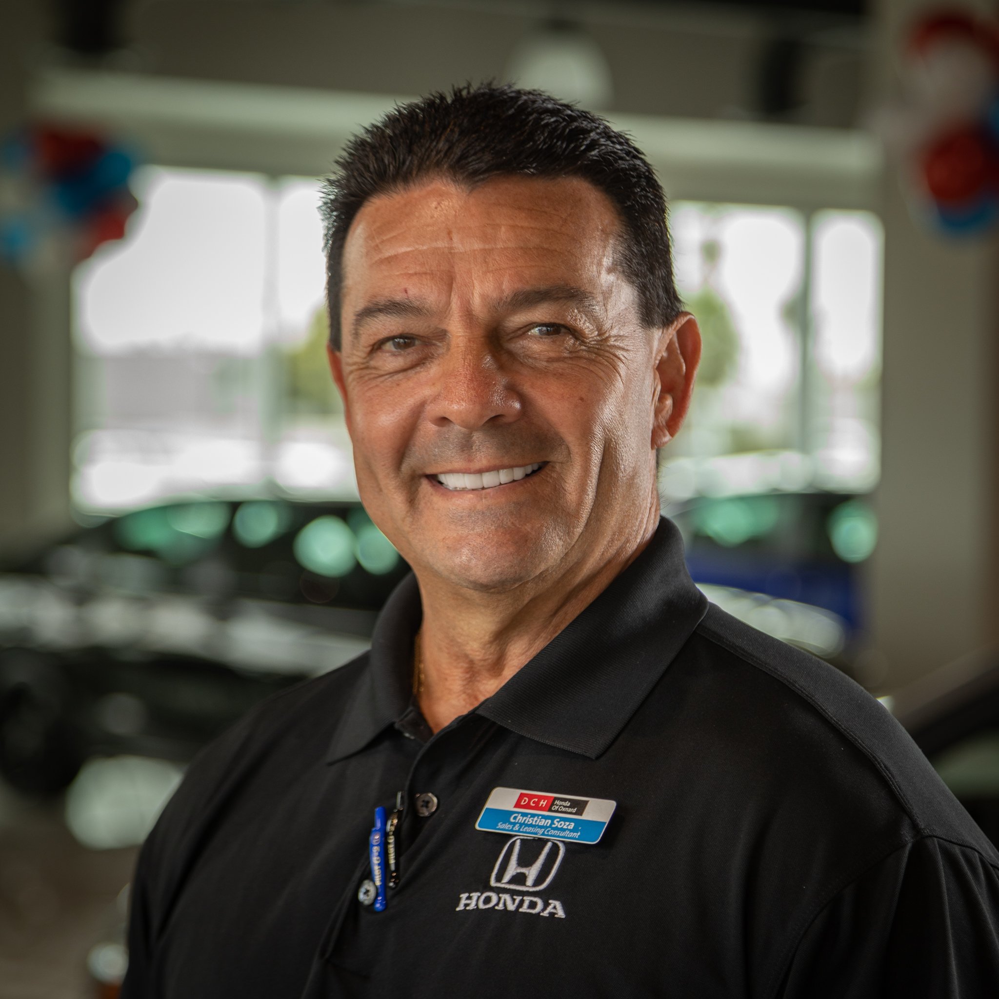 A smiling man wearing a black polo shirt with Honda logo and a name tag, standing inside a car dealership with cars and large windows in the background.