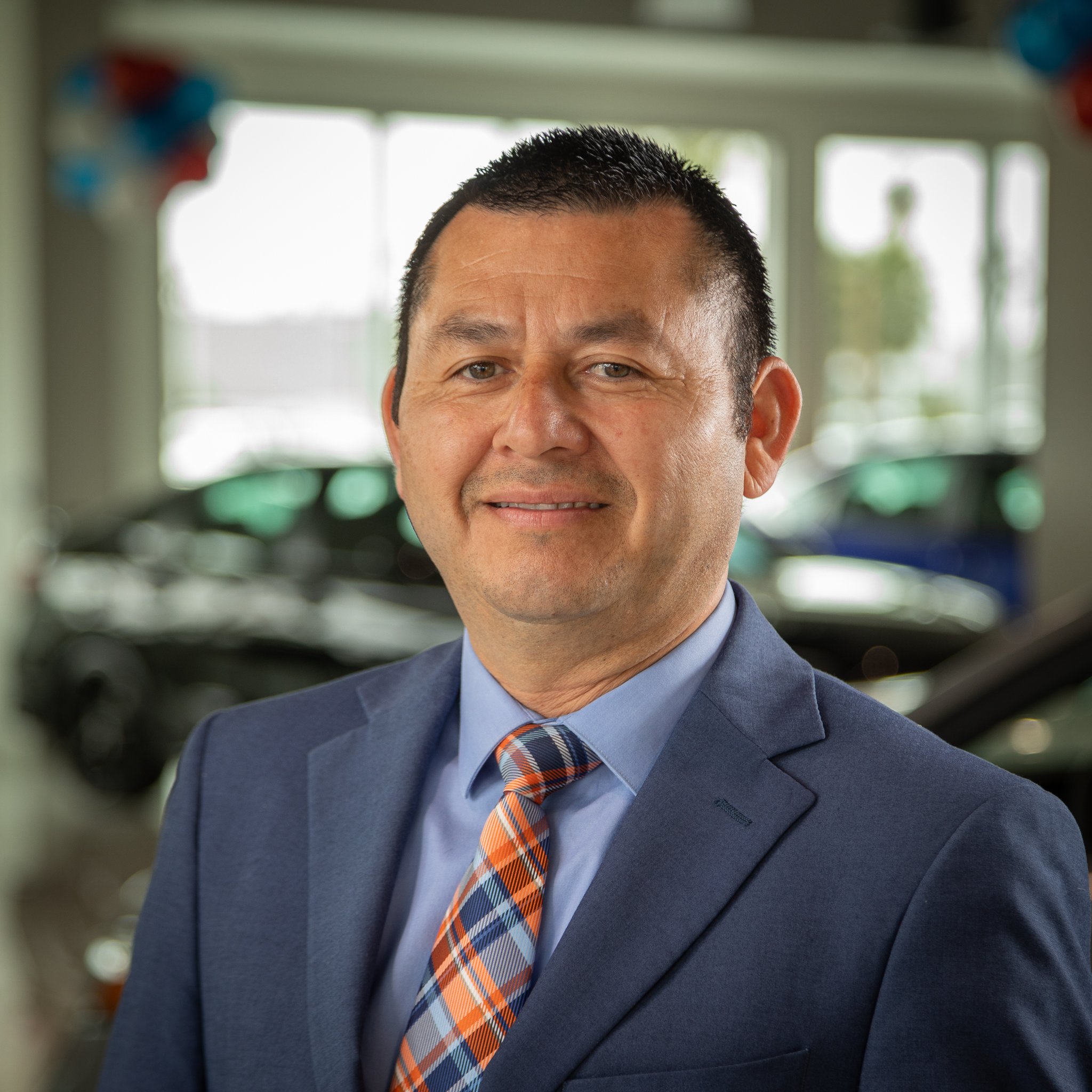 A man in a blue suit and multicolored tie standing inside a car dealership.