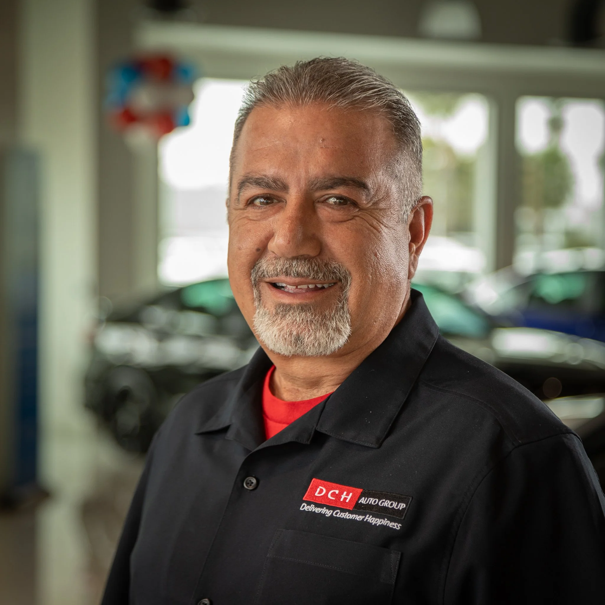 A middle-aged man with gray hair and a beard, smiling, dressed in a black uniform with a red shirt underneath, standing inside a car dealership.