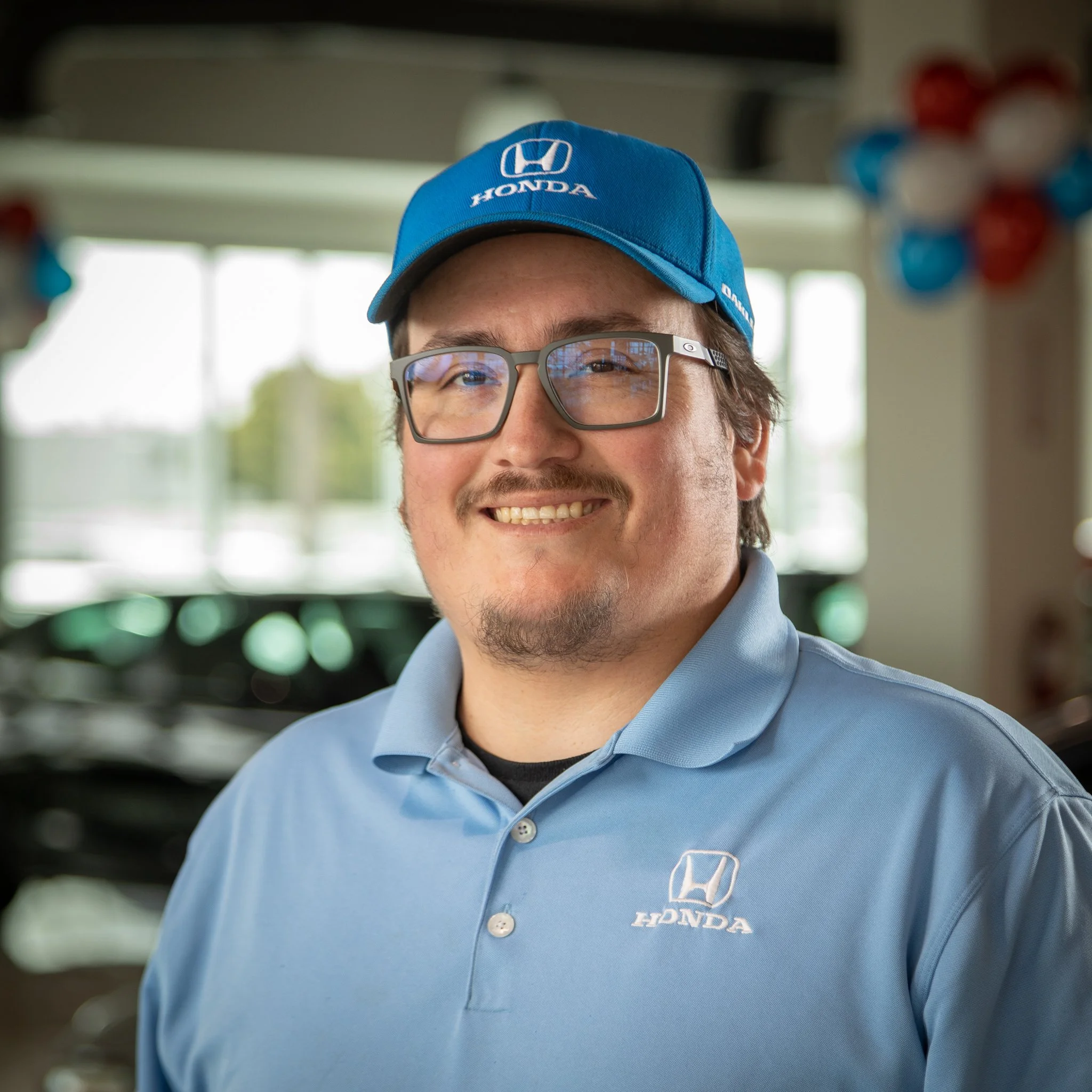 A man smiling in a Honda dealership, wearing a blue Honda cap, glasses, and a light blue Honda polo shirt.