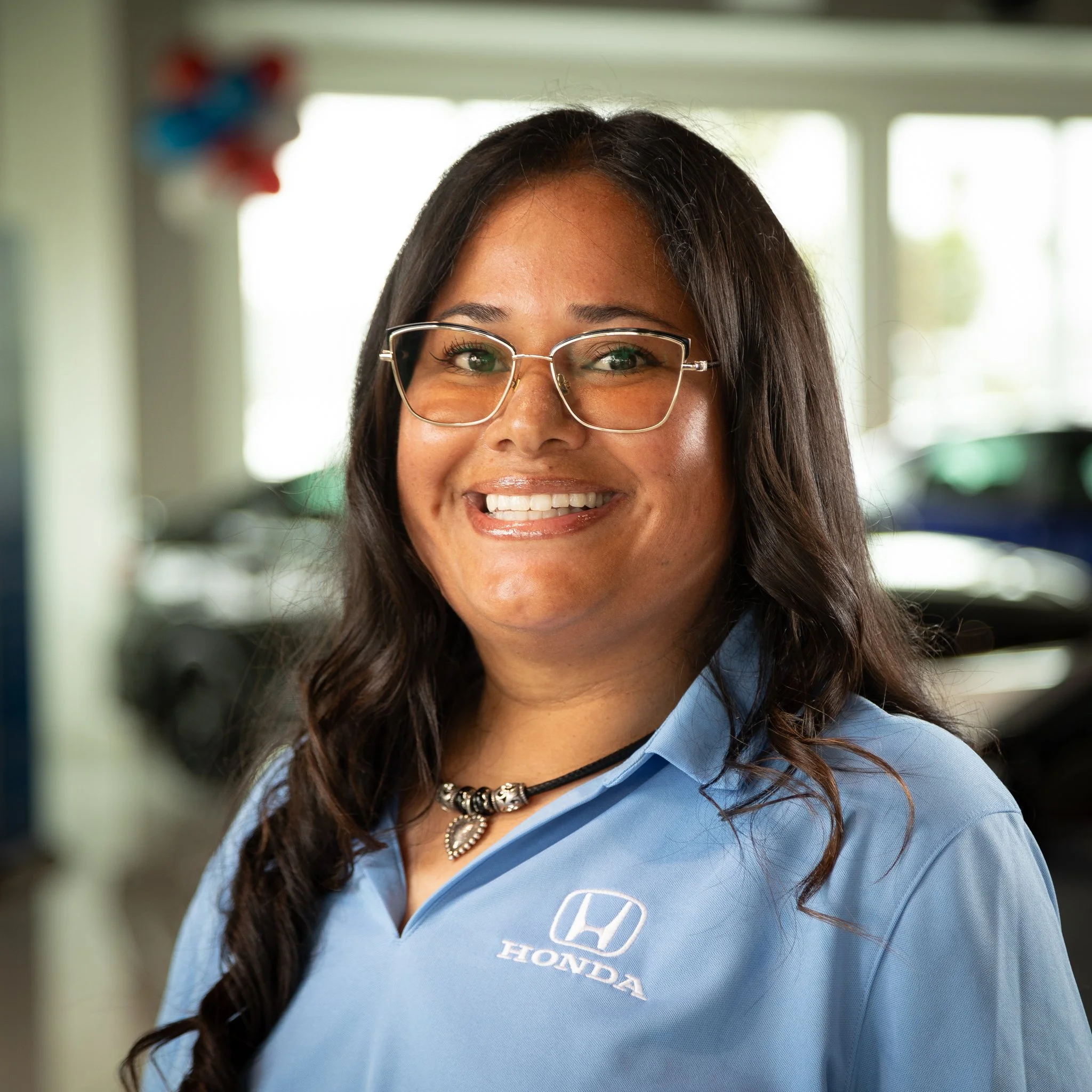 A woman with long dark hair wearing glasses and a blue Honda polo shirt, smiling indoors with cars in the background.