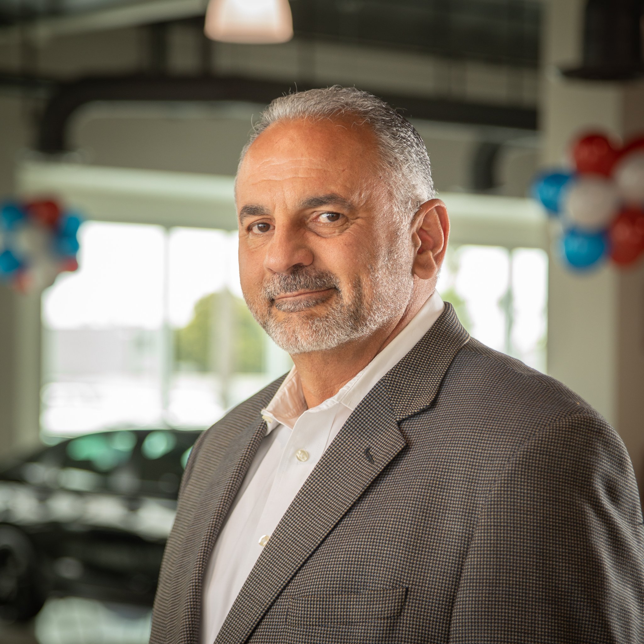A middle-aged man with a gray beard and short gray hair, wearing a brown checkered blazer and a white shirt, standing indoors with a neutral expression, blurred background with windows and balloons.