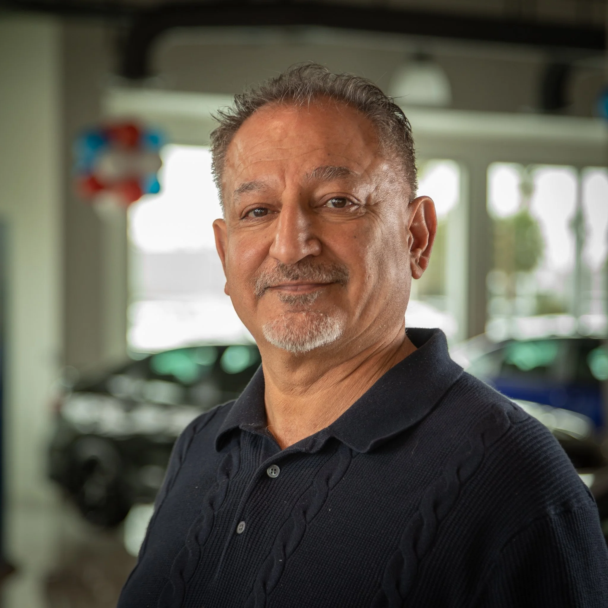A middle-aged man with salt and pepper hair and a goatee, wearing a dark polo shirt, standing inside a car dealership. Blurred cars are visible in the background.