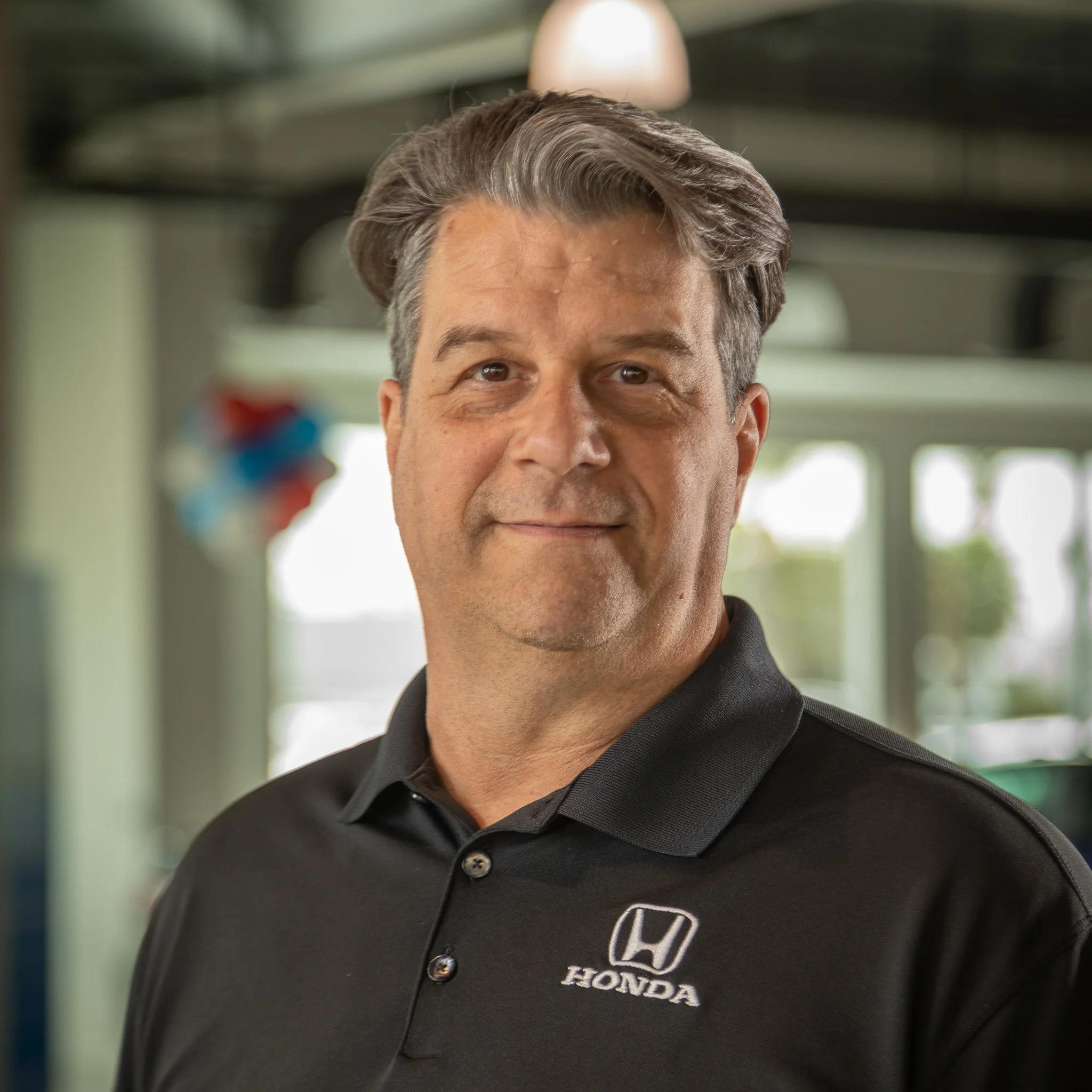 A middle-aged man with gray hair and a slight smile, wearing a black Honda polo shirt, standing inside a showroom or service area with a blurred background.