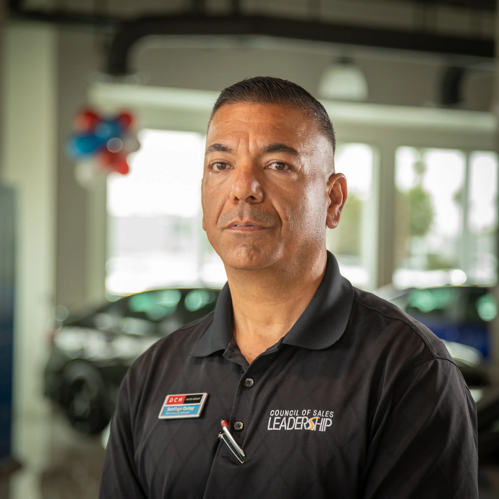 A man wearing a black polo shirt with the logo 'Council of Sales Leadership' and a name badge, standing inside a car dealership with cars visible in the background.