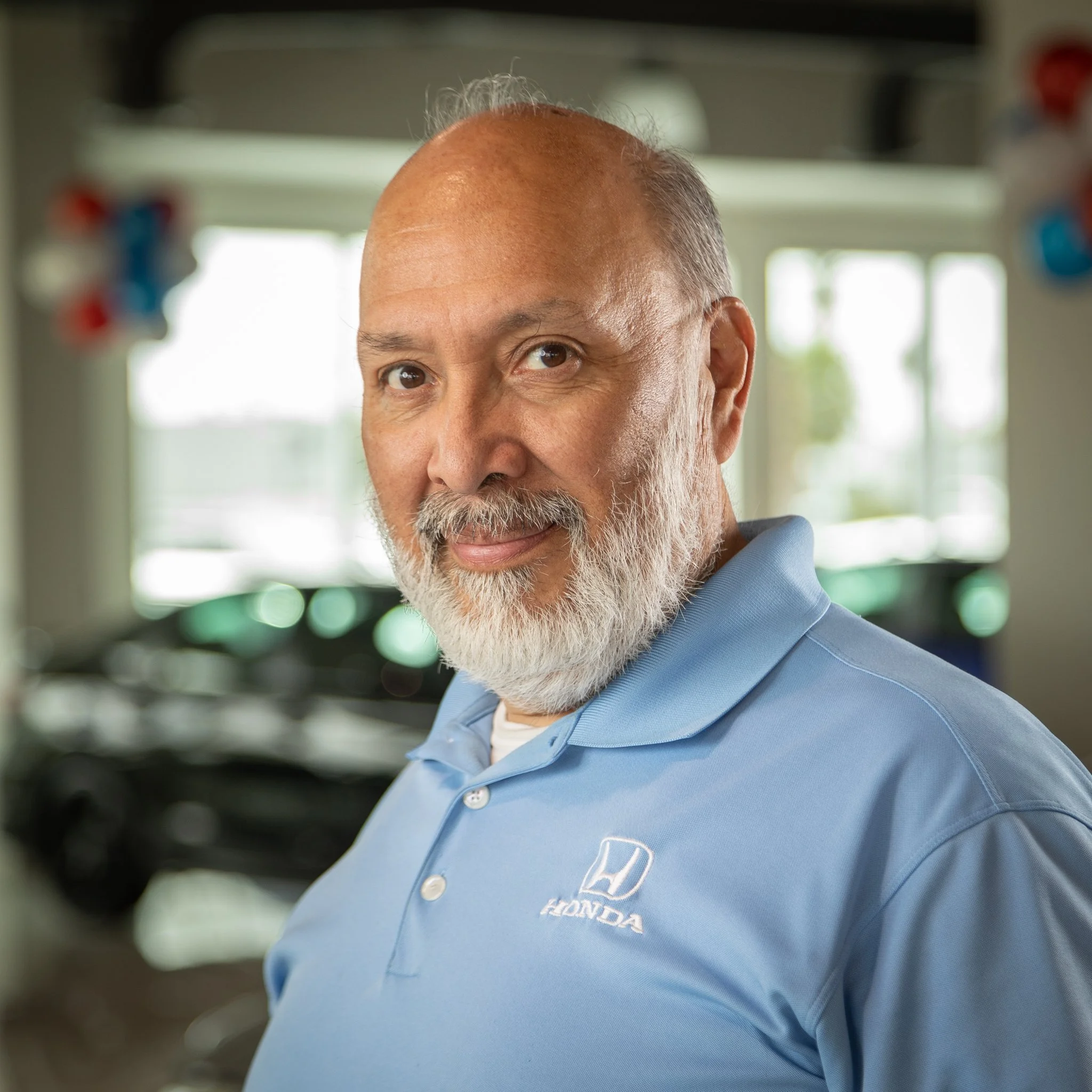 A middle-aged man with a beard and mustache wearing a light blue Honda work shirt, standing inside a car dealership, looking at the camera with a confident expression.