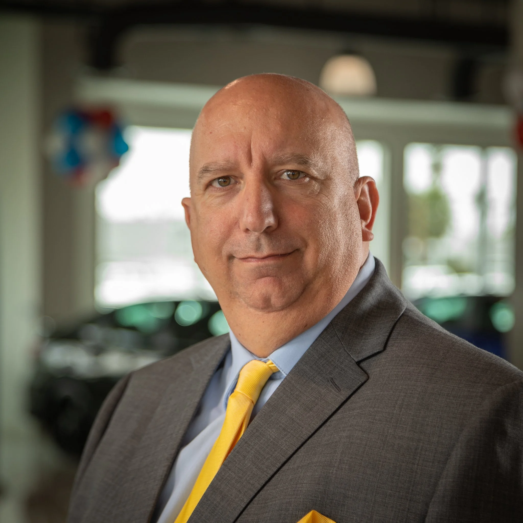 A bald man in a gray suit with a yellow tie, standing in an auto dealership showroom with cars visible in the blurred background.