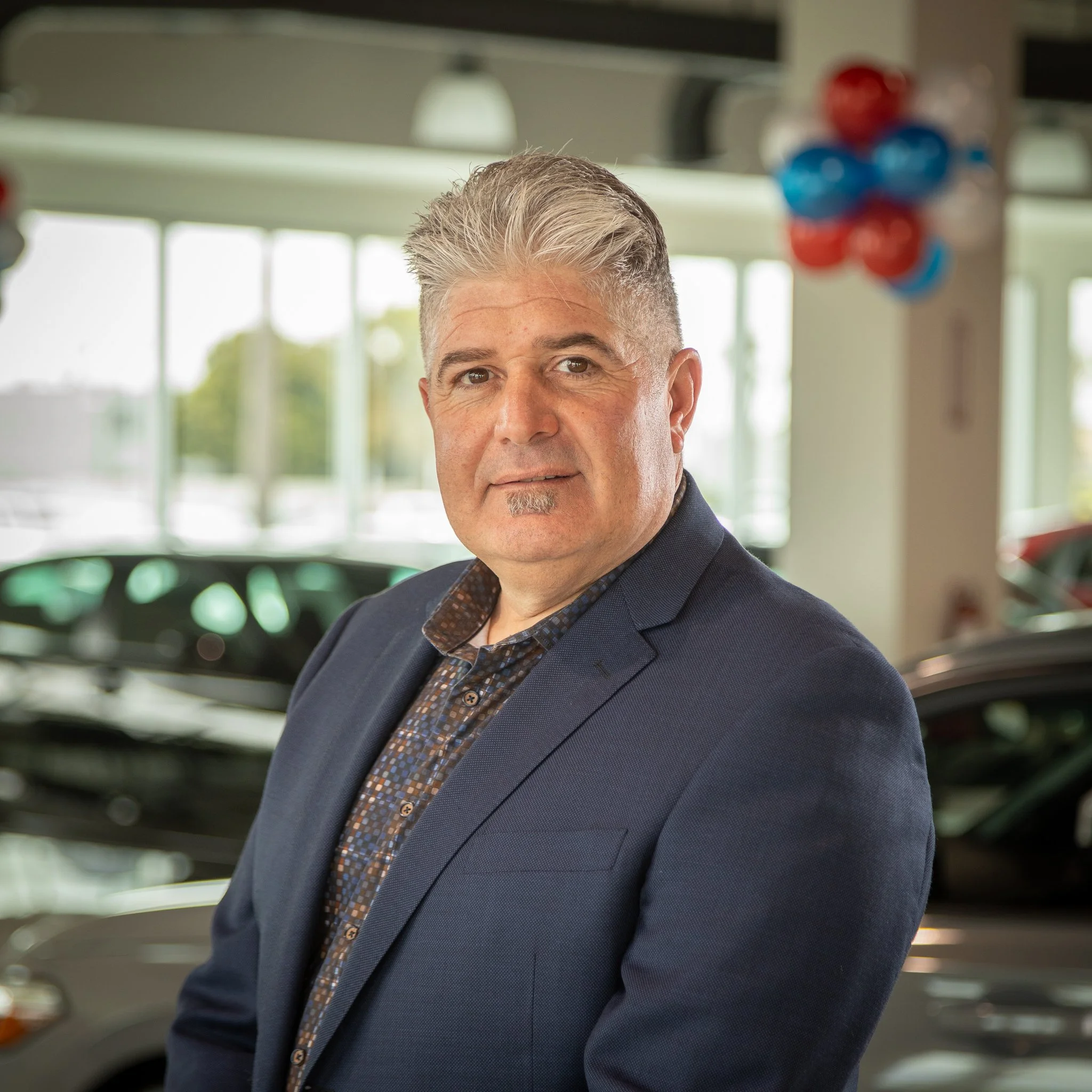 A middle-aged man with short gray hair and a goatee, dressed in a navy blazer and patterned shirt, standing inside car dealership with cars in the background and red, white, and blue balloons hanging up.