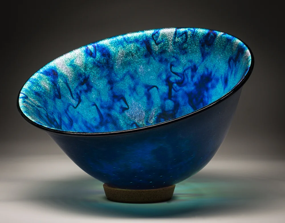 “Cosmic Powers” 7”H X 9.5” W This vessel began as 2 pieces of sheet glass cut into 16” circles and fused in a kiln to create a single flat disc. The “slumping” process is where the glass is placed on a mold and warmed in the kiln until the glass takes the shape of the mold. To gain this shape is a 2 step processes using graduated sized molds to attain the shape. The piece is purposely slanted to allow for seeing the center of the vessel