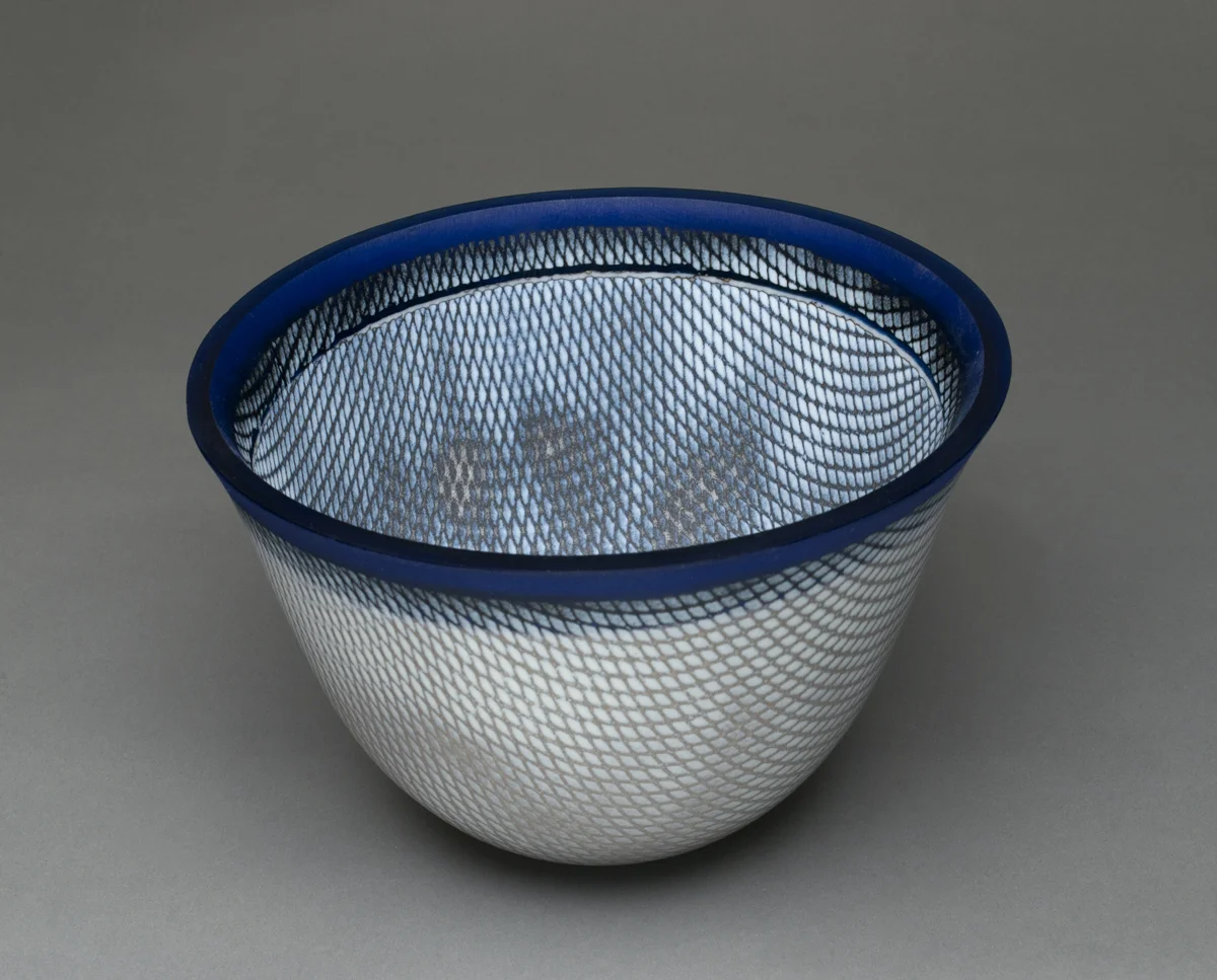 “Shades of Blue” 7”H X 9.5” W This vessel began with cutting a 16” ring of sheet glass (blue) and 14” circle of white glass. A stencil was placed on one side and glass powder was sifted onto it. The piece was fused in a kiln and then the other side was stenciled. The “slumping” process is where the glass is placed on a mold and warmed in the kiln until the glass takes the shape of the mold. To gain this shape is a 3 step processes using graduated sized molds to attain the shape. The edge is ground and polished using a grinding wheel and water.