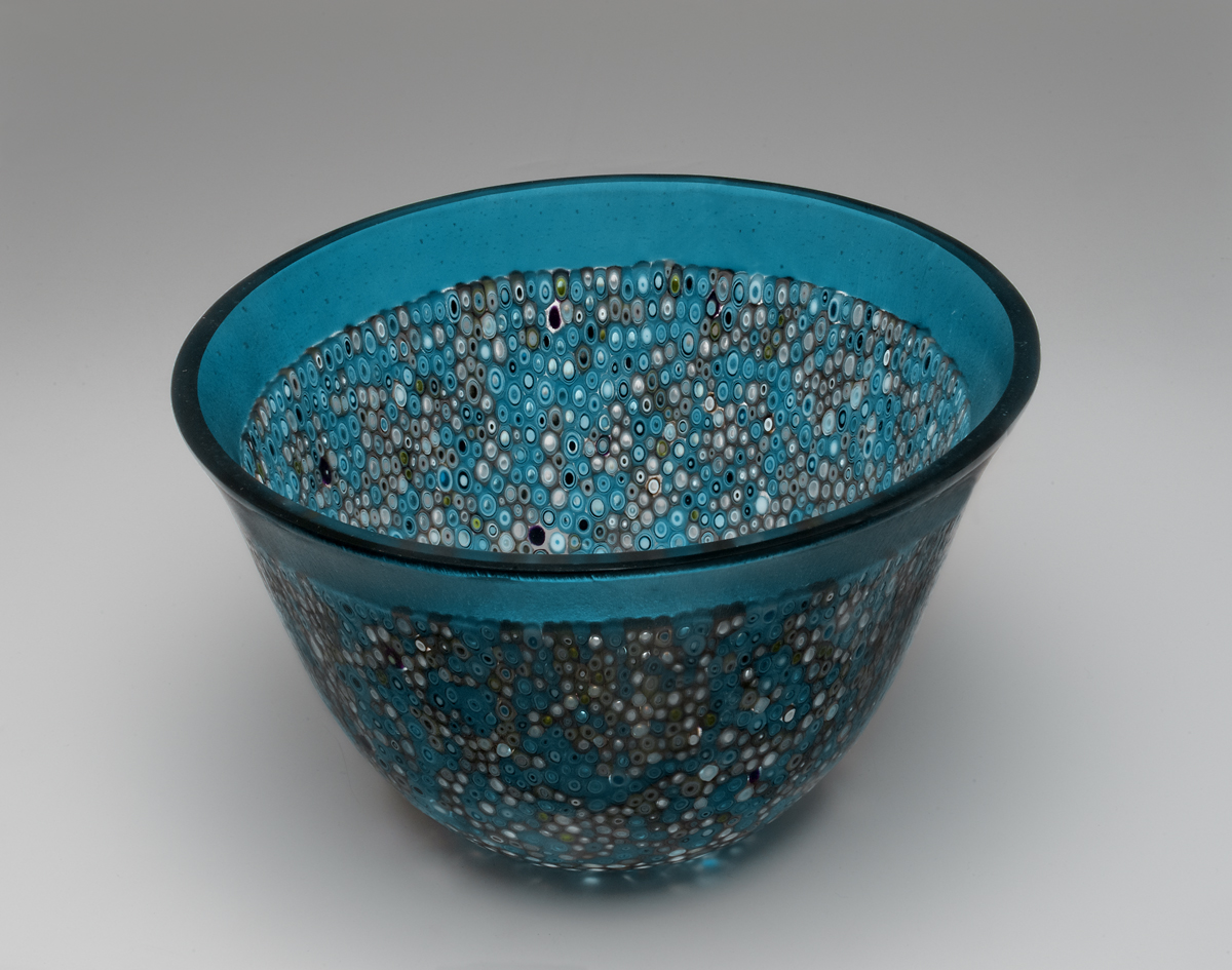 “Blue Bubbles” 7”H X 9.5” W This vessel began with many cutting a 16” circle, then a 2” outer ring was cut and pre-made murrine were placed within the ring. All of the components were fired to create a 16” flat circle. The “slumping” process is where the glass is placed on a mold and warmed in the kiln until the glass takes the shape of the mold. To gain this shape is a 3 step processes using graduated sized molds to attain the shape. The edge is ground and polished using a grinding wheel and water.