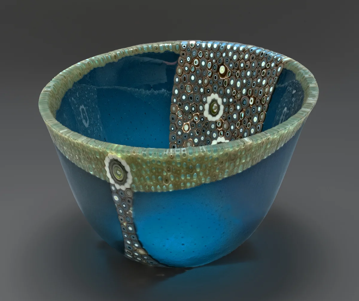 This vessel began by cutting a 14” circle and offsetting it to allow the addition of pre-made murrine to be added along the rim and throughout the center. It was then fired to make one flat 16” circle The “slumping” process is where the glass is placed on a mold and warmed in the kiln until the glass takes the shape of the mold. To gain this shape is a 3 step processes using graduated sized molds to attain the shape. The edge is ground and polished using a grinding wheel and water