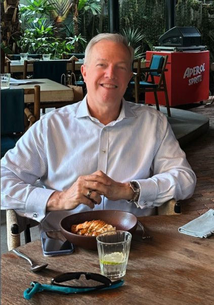 US Ambassador Ted Osius sitting at a restaurant table with a bowl of food, a glass of water, and a smartphone, smiling with a background of lush green plants and restaurant decor.