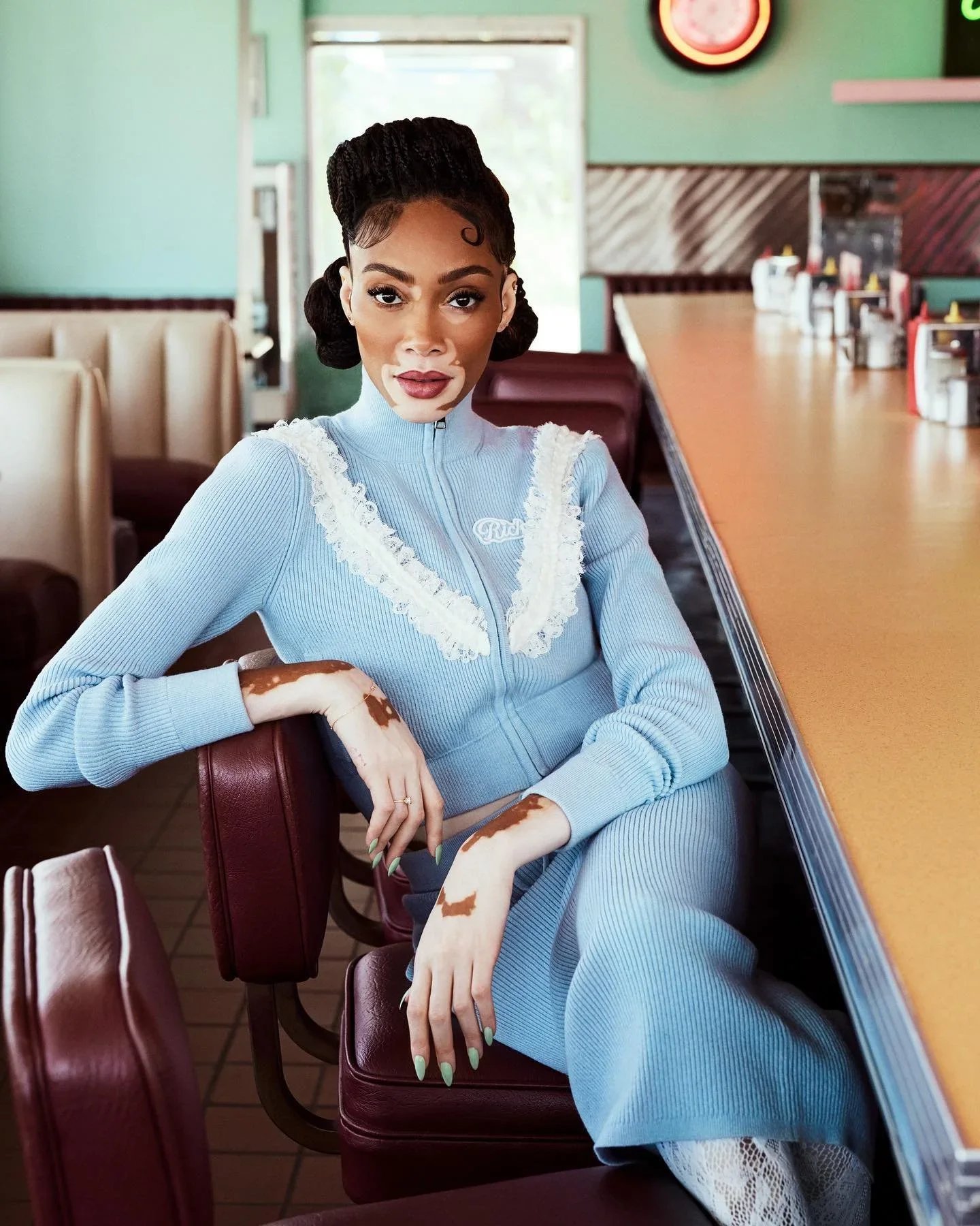Winnie Harlow seated in a retro diner wearing a pale blue knit set and mint green nails.