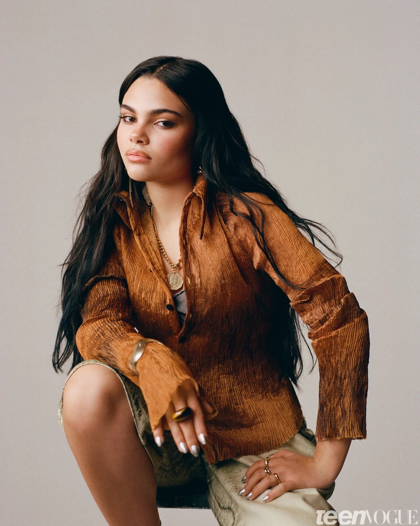 Studio portrait of Ariana Greenblatt wearing a textured bronze-brown button-down shirt and layered gold jewelry. Metallic silver nails complement the editorial styling.