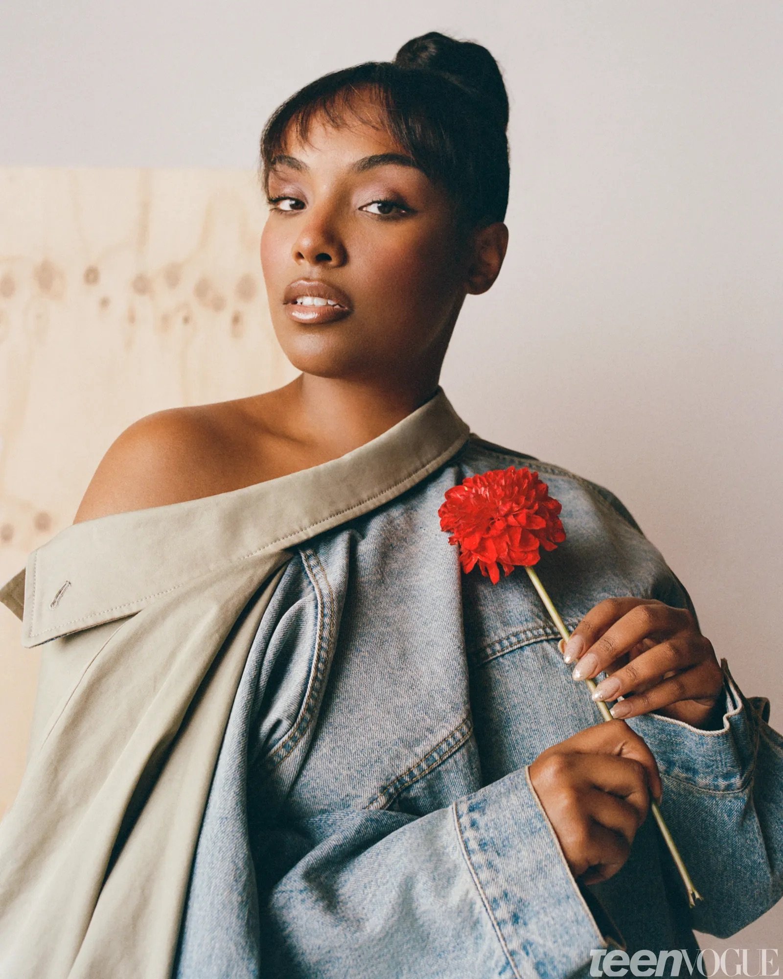 Studio portrait of Aida Osman wearing an oversized denim jacket draped off one shoulder while holding a bright red flower. Soft lighting and neutral tones create a modern editorial look.