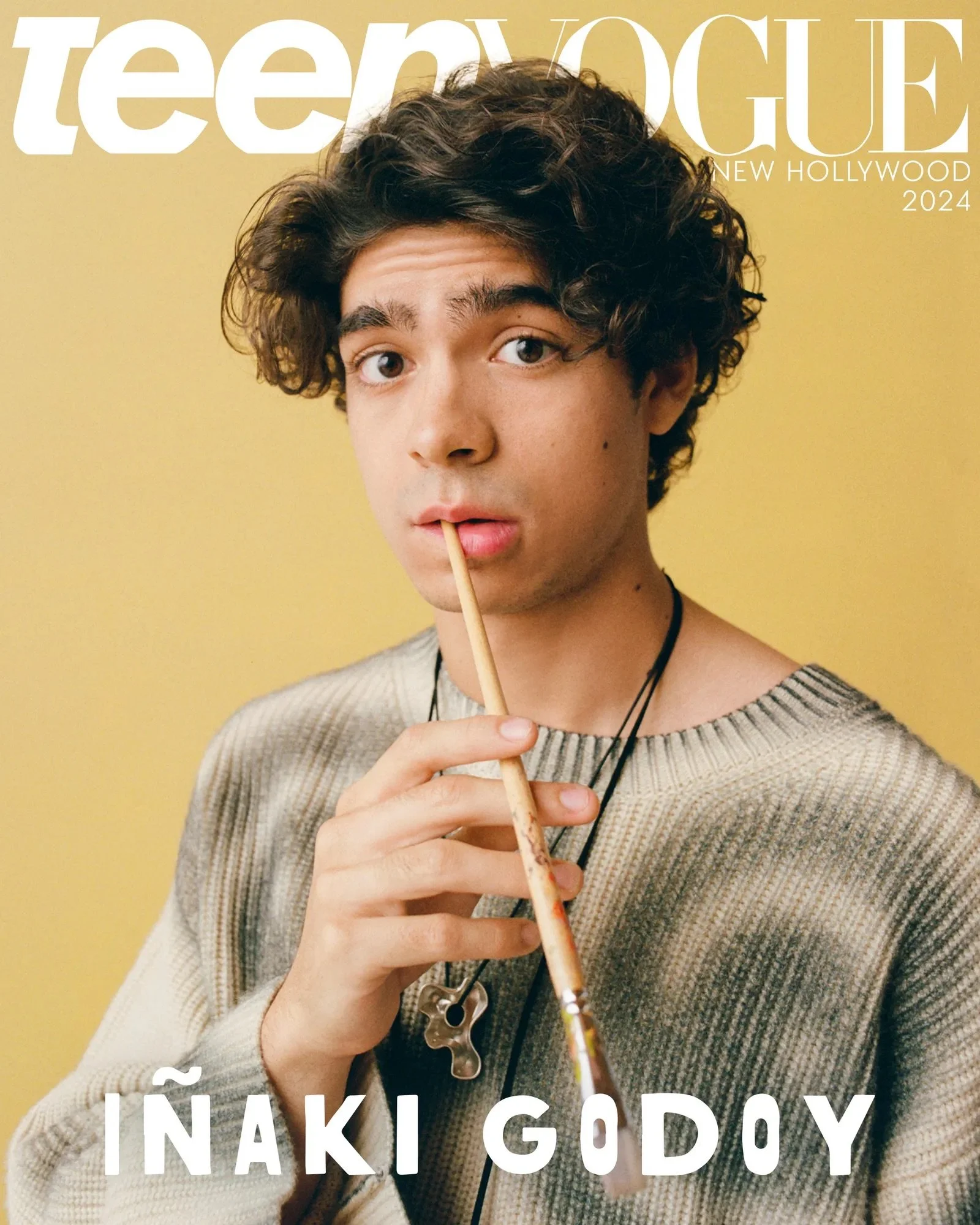 Studio portrait of Iñaki Godoy holding a paintbrush to his lips while wearing a textured knit sweater and pendant necklace against a yellow background.