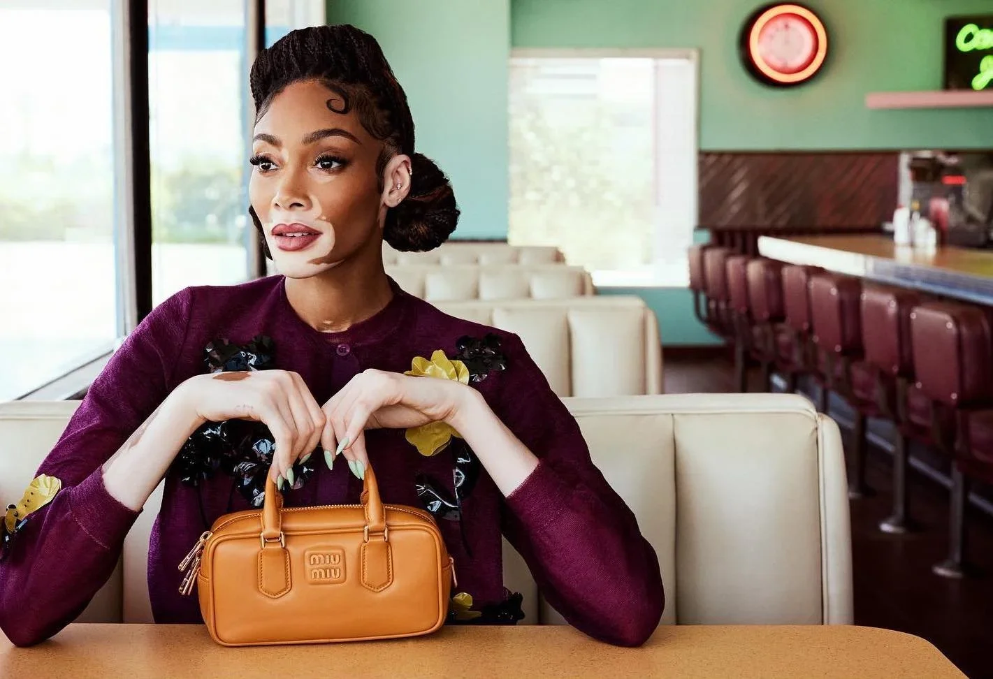 Winnie Harlow seated in a diner booth wearing a burgundy outfit and holding a tan Miu Miu handbag.