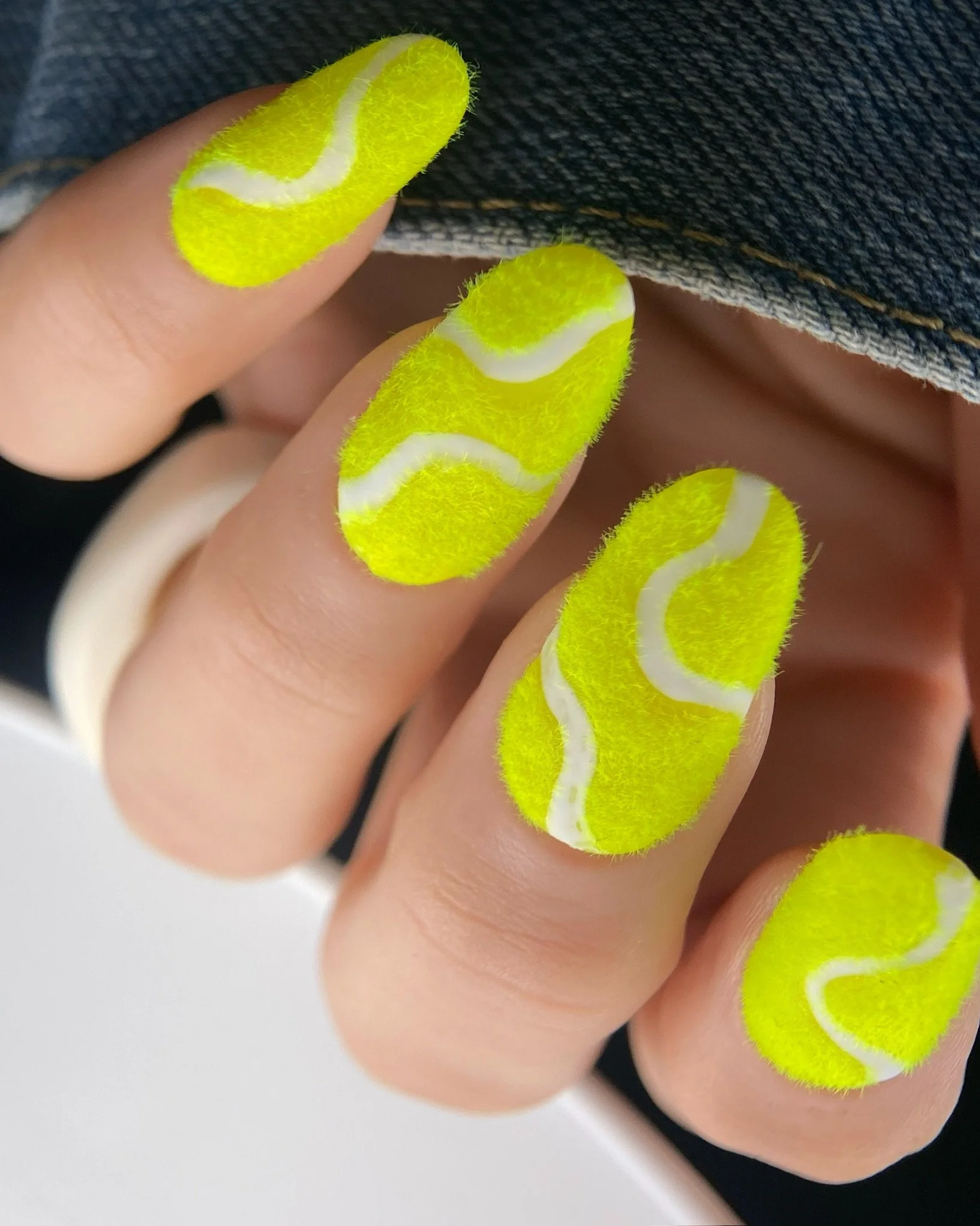 Close-up of fuzzy tennis ball nail art with bright neon yellow flocking powder and white curved seam lines.