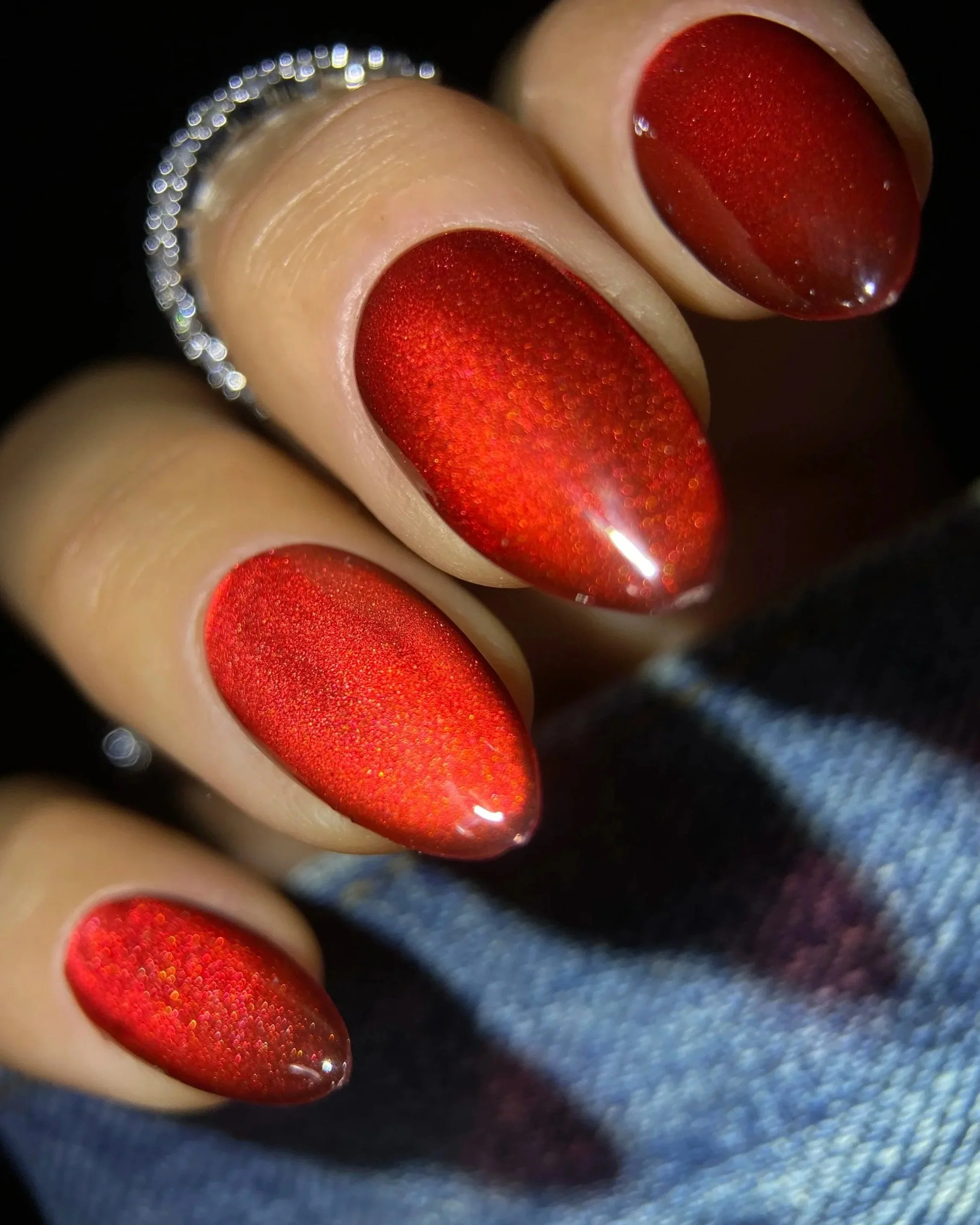 Close-up of Double Rhythm Gemstone Glow MC1327, a jewel-toned ruby red magnet gel with a luminous velvet magnetic finish on an almond-shaped nail tip photographed against denim.