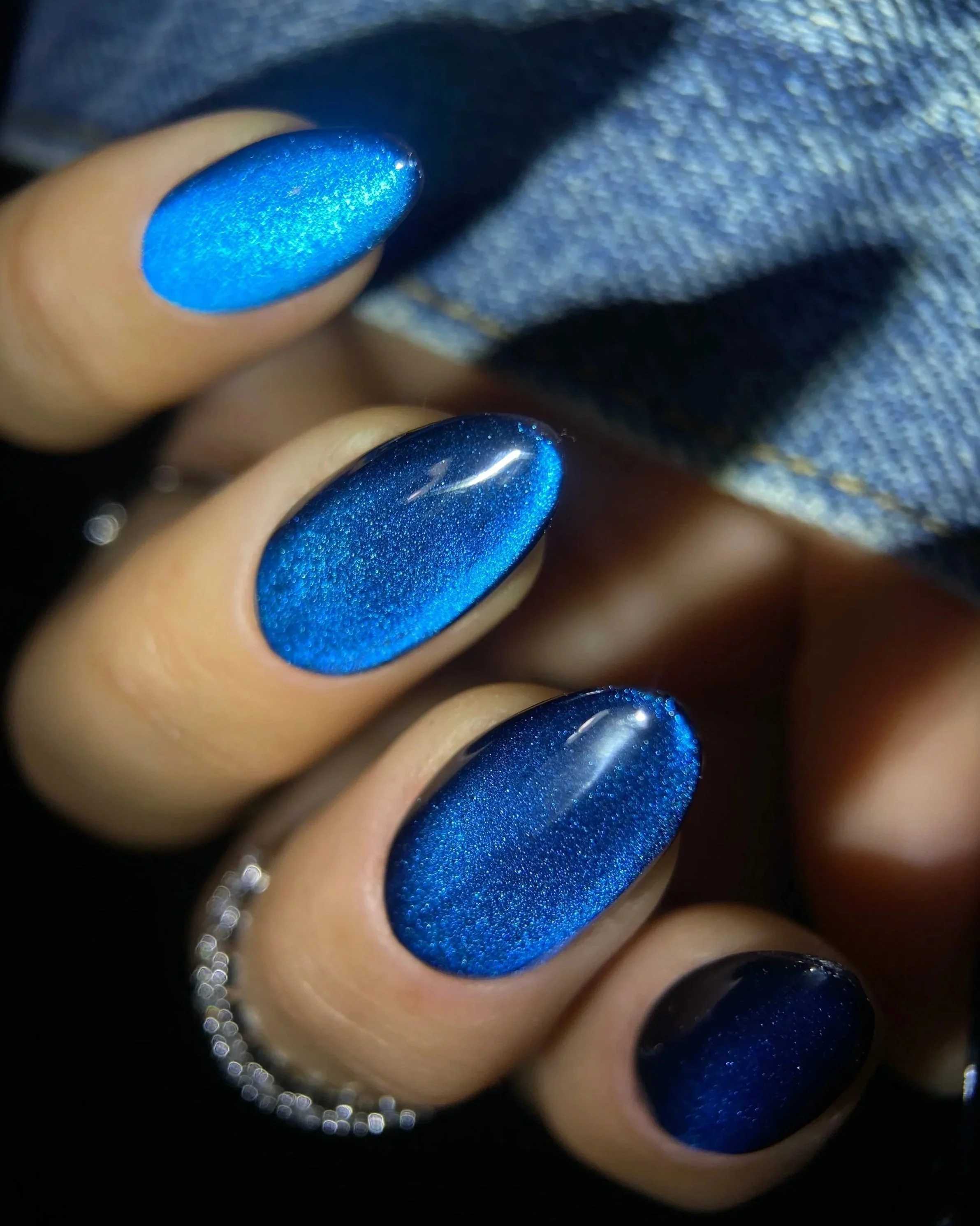Close-up of Double Rhythm Gemstone Glow MC1312, a rich sapphire-blue magnet gel with fine magnetic particles creating a bright velvety cat-eye effect on an almond-shaped nail tip against denim.