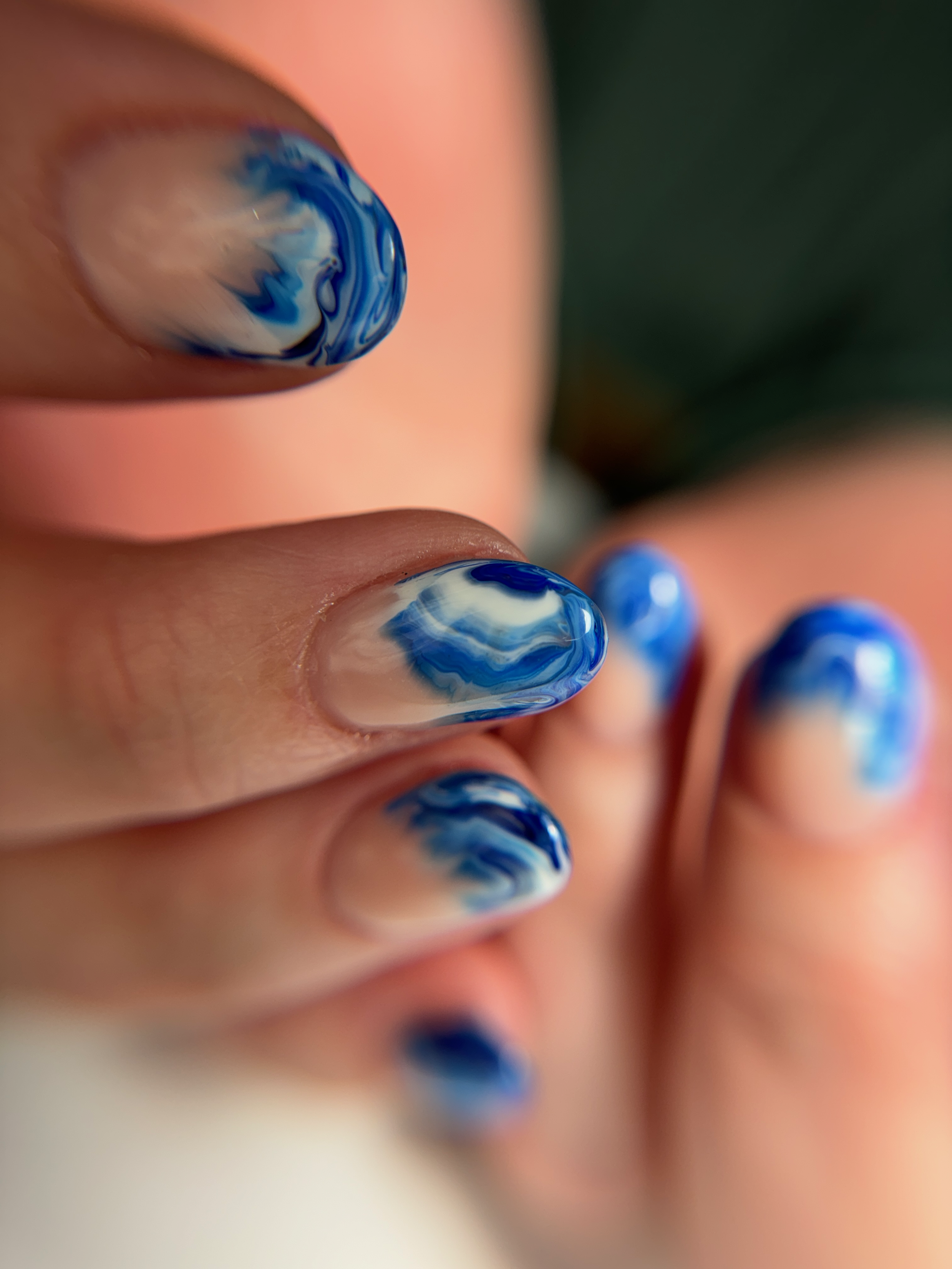 Close-up of blue and white ocean-inspired marble nail art created with blooming gel. Swirls of cobalt, sky blue, and white flow across a sheer nude base, creating a fluid wave-like effect reminiscent of crashing surf and seafoam along the shoreline.