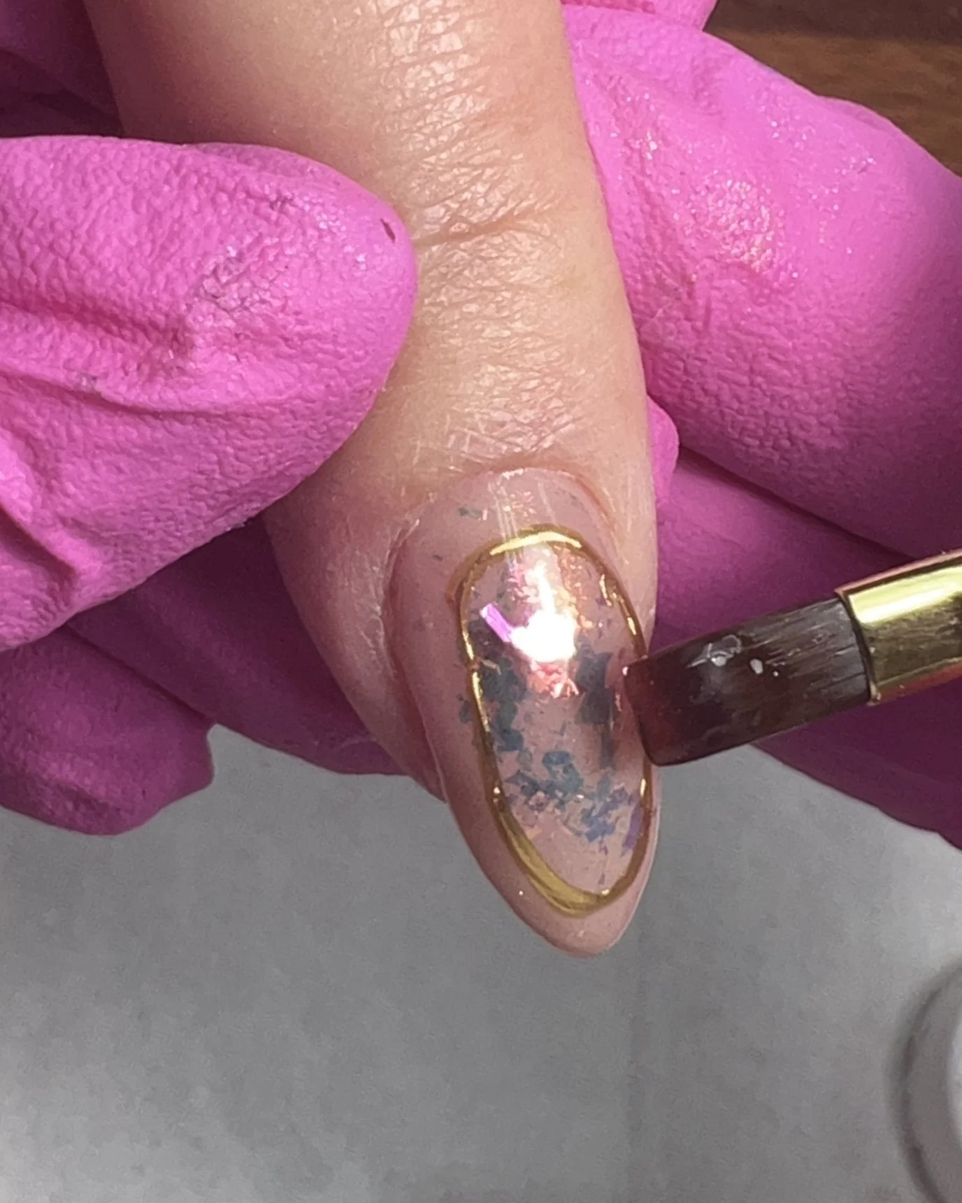 A brush applies clear builder gel over a brassy gold chrome frame and encapsulated opal flakes to seal the design before top coating.